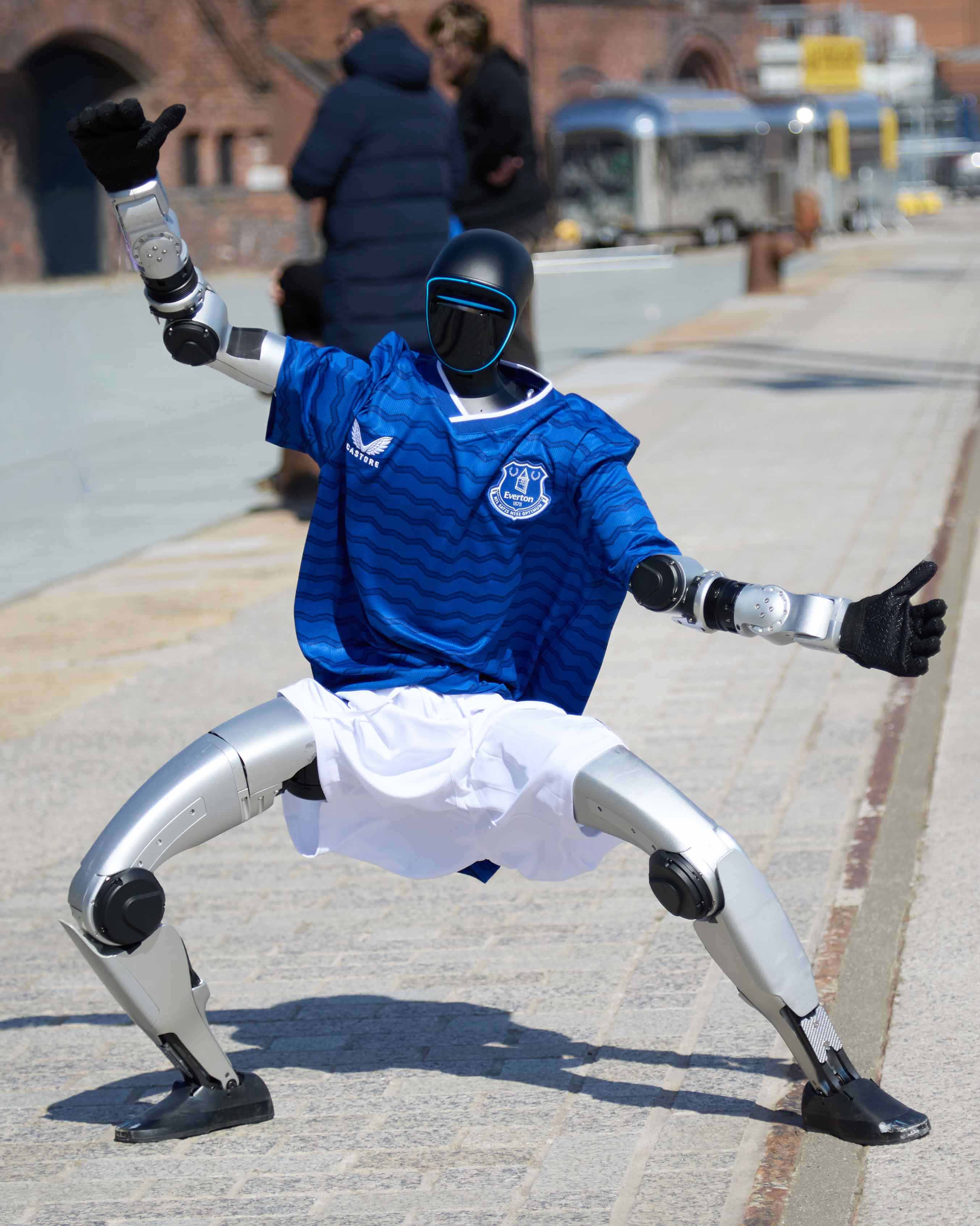 Humanoid robot wearing an Everton football kit performs moves on a walkway in front of buildings