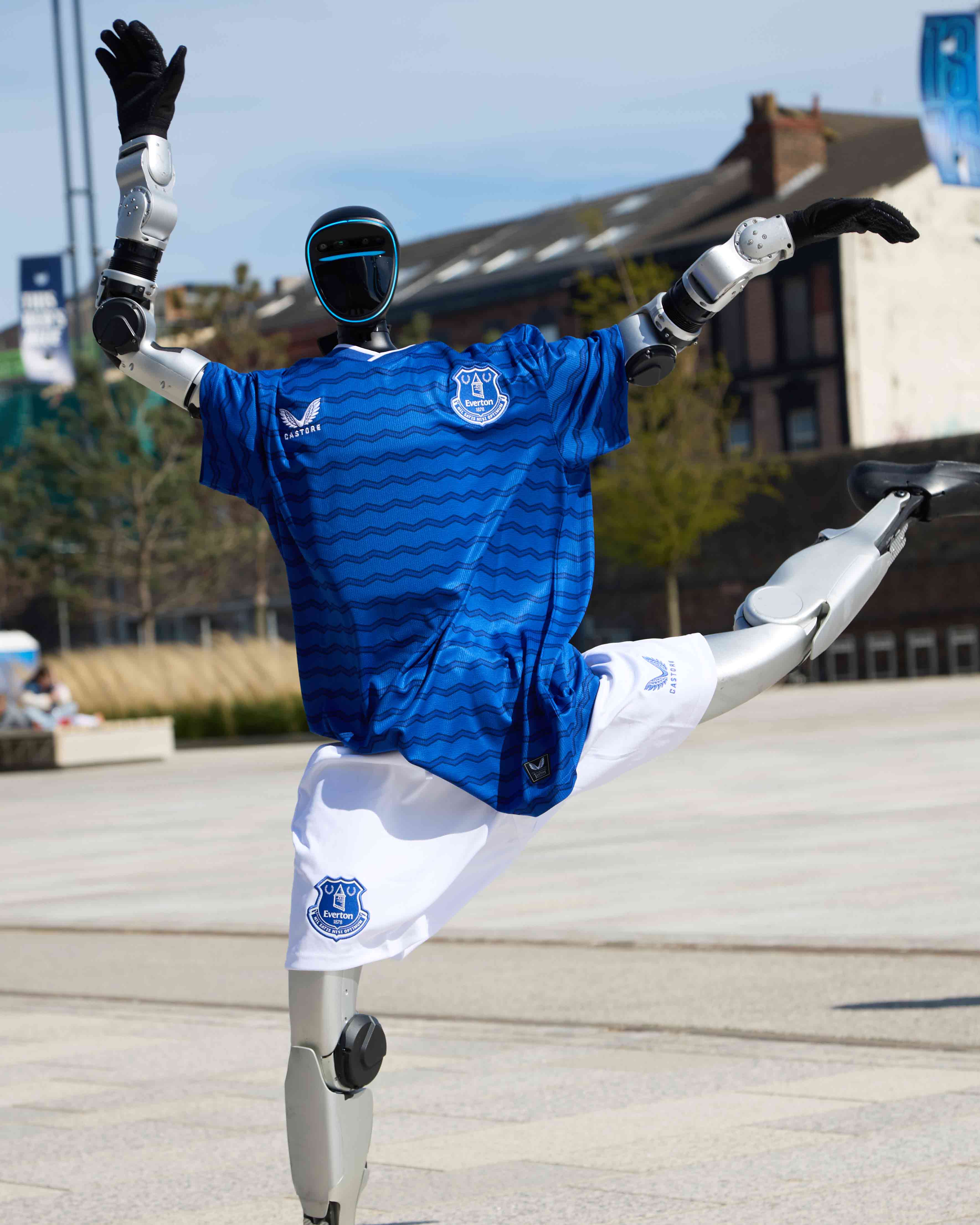 Humanoid robot wearing an Everton football kit performs a one legged stand on a walkway in front of buildings