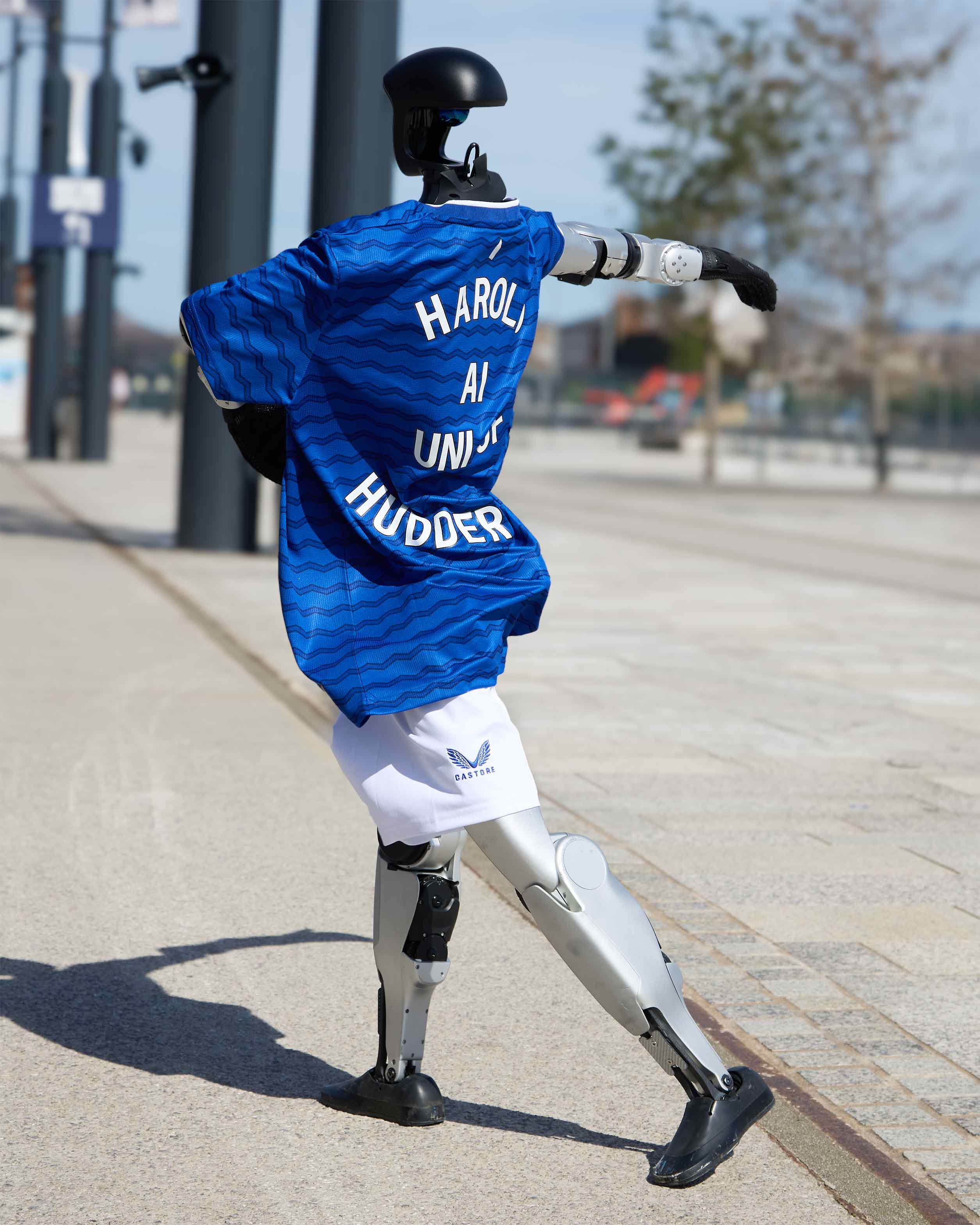 Humanoid robot wearing an Everton football kit dances on a walkway in front of buildings