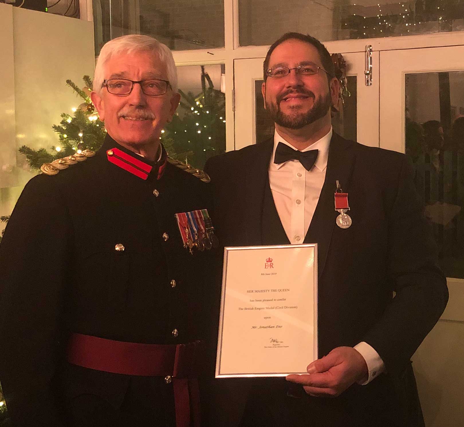 Jon Eno wearing tuxedo holding his certificate for his British Empire Medal award