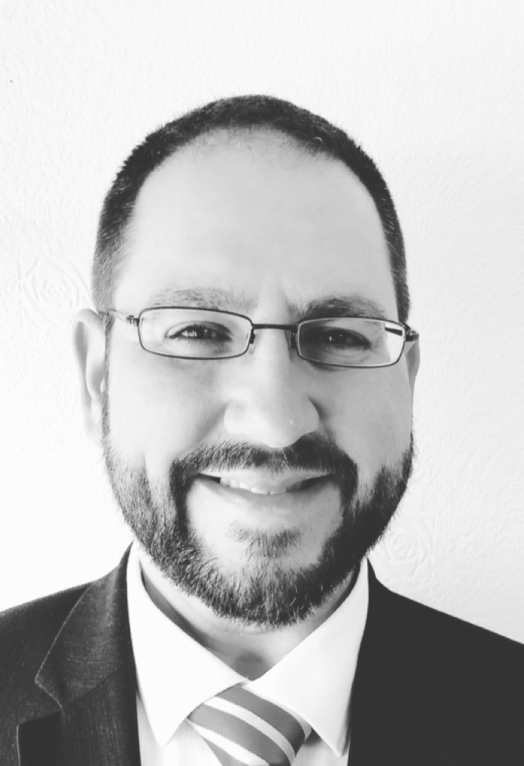 Black and white close up shot of Jon Eno smiling, wearing a suit and glasses.