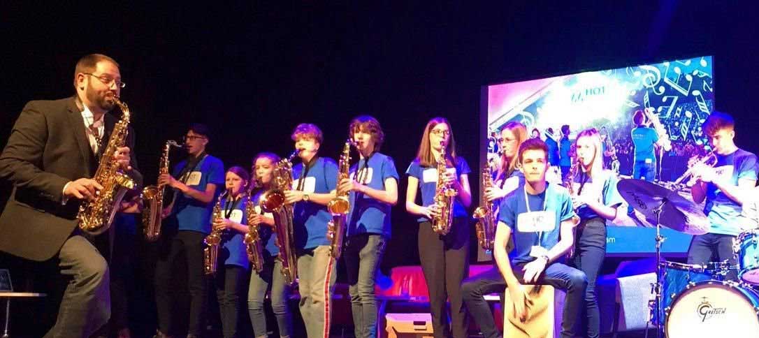 Jon Eno playing with Saxophonix infront of a group of students also playing musical instruments.