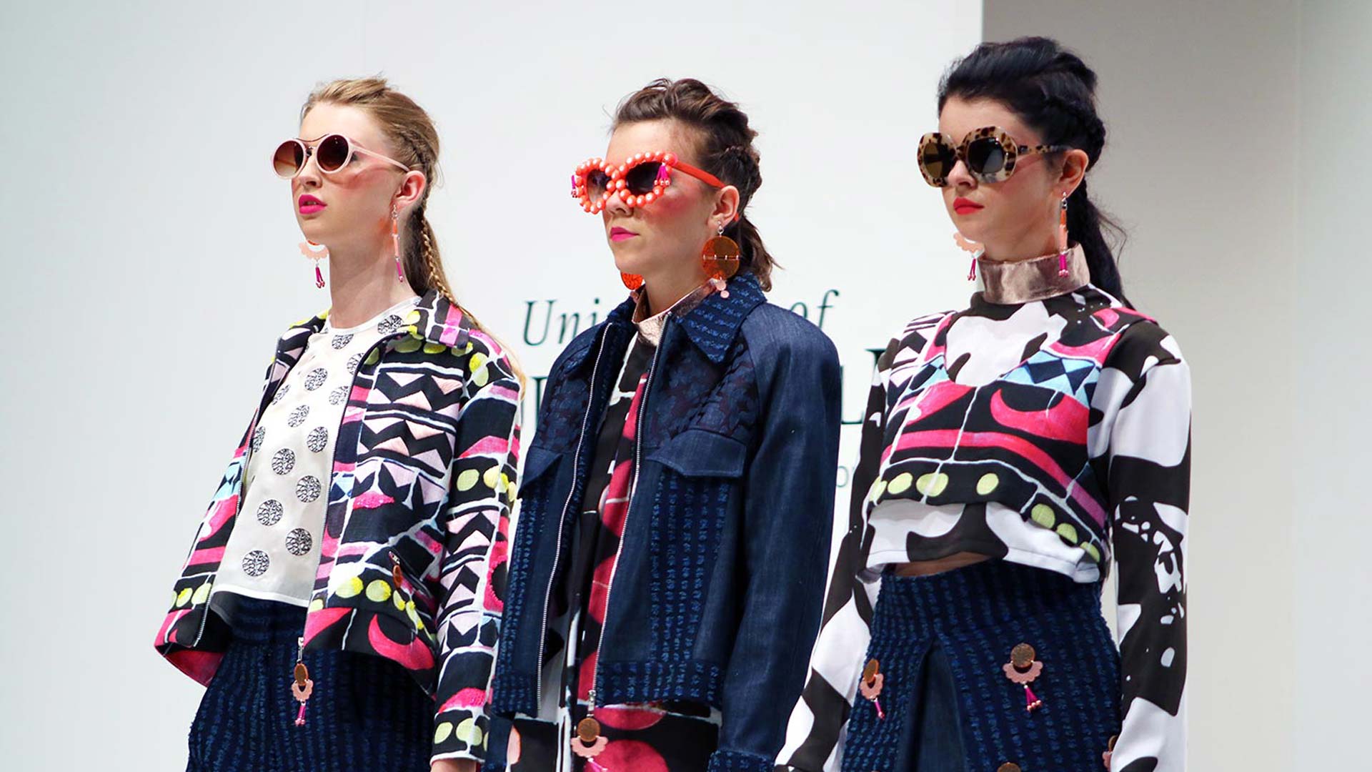 Fashion design image of 3 models posing side by side. Model 1 wears sunglasses, patterned jacket and blue jeans. Model 2 wears denim jacket with a patterned dress. Model 3 wears a long sleeved patterned top and blue skirt.