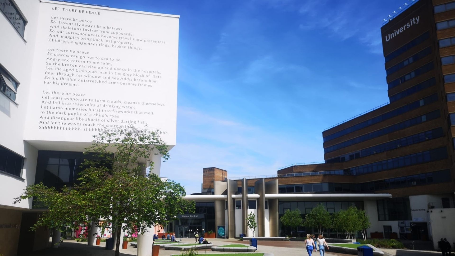 A large white building with a poem written on its side, redbrick building and silver building surrounding a paved area