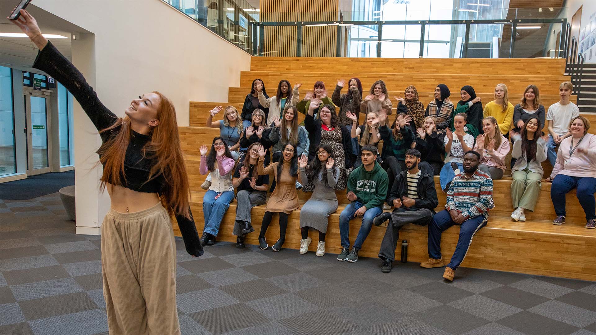 A person is taking a selfie with a group seated on wooden steps inside a modern building. The group is posing and waving. The atmosphere appears cheerful and casual.