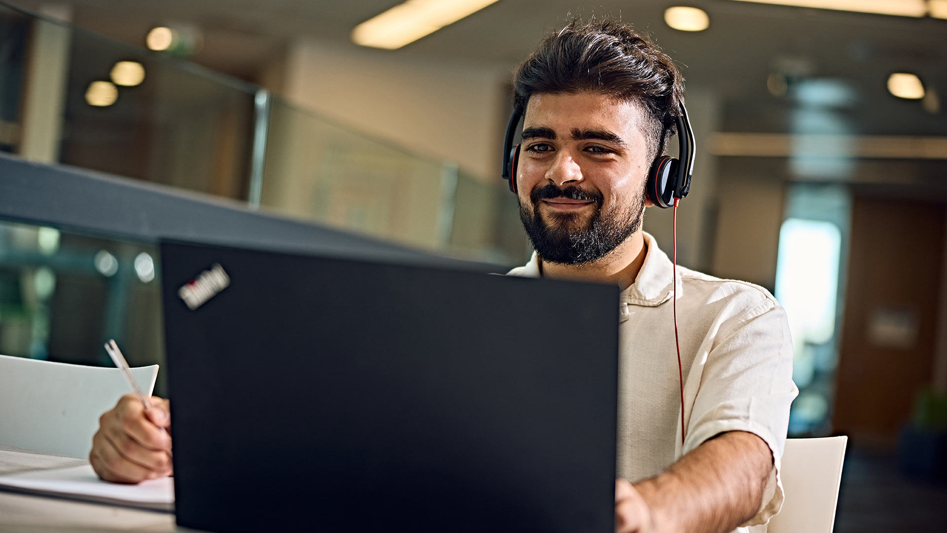 A smiling distance learning student wearing a headset, writing notes and working on a laptop.