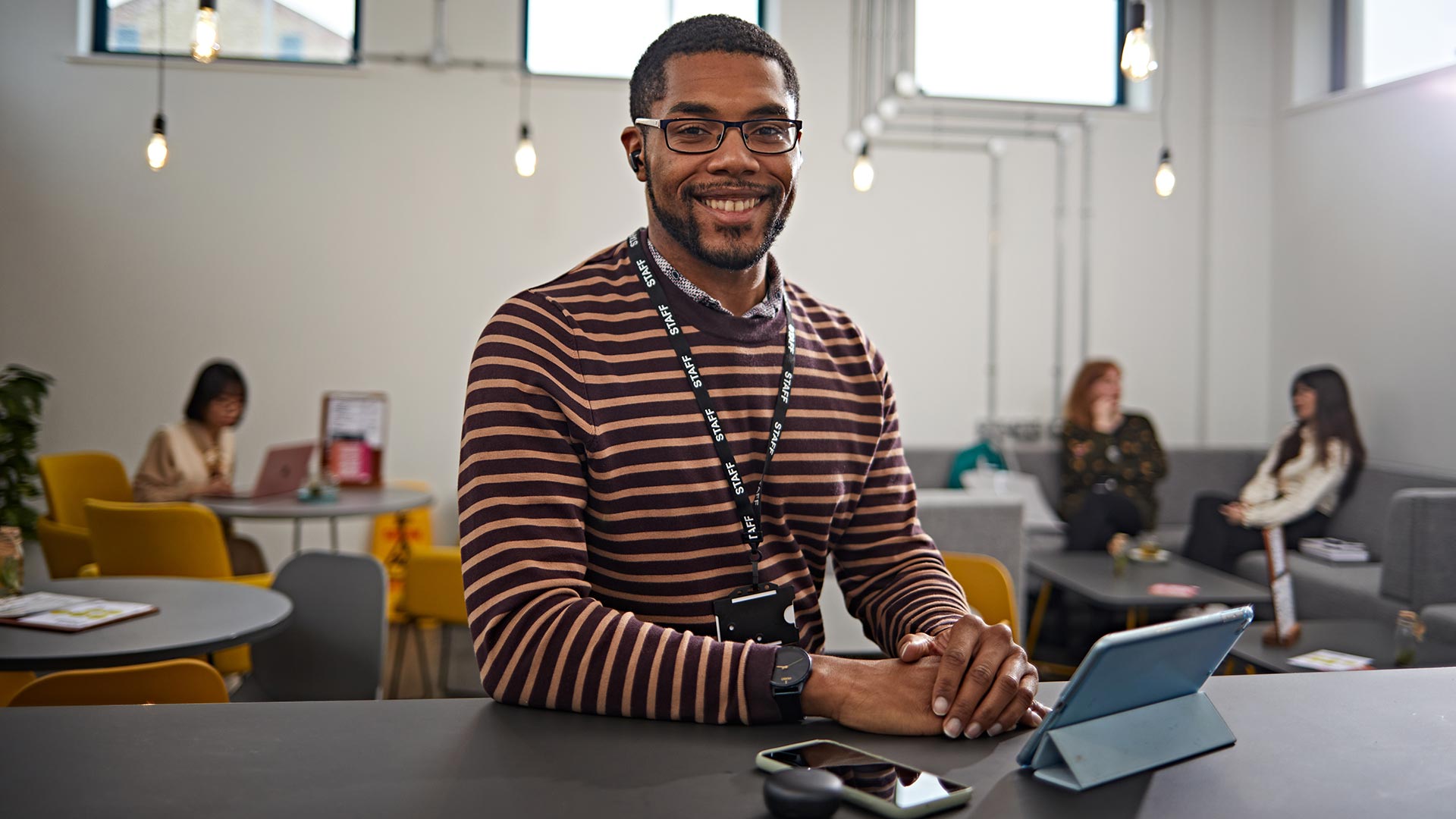 A smiling student sat in a café, with a tablet device, mobile and earbud case on the table.