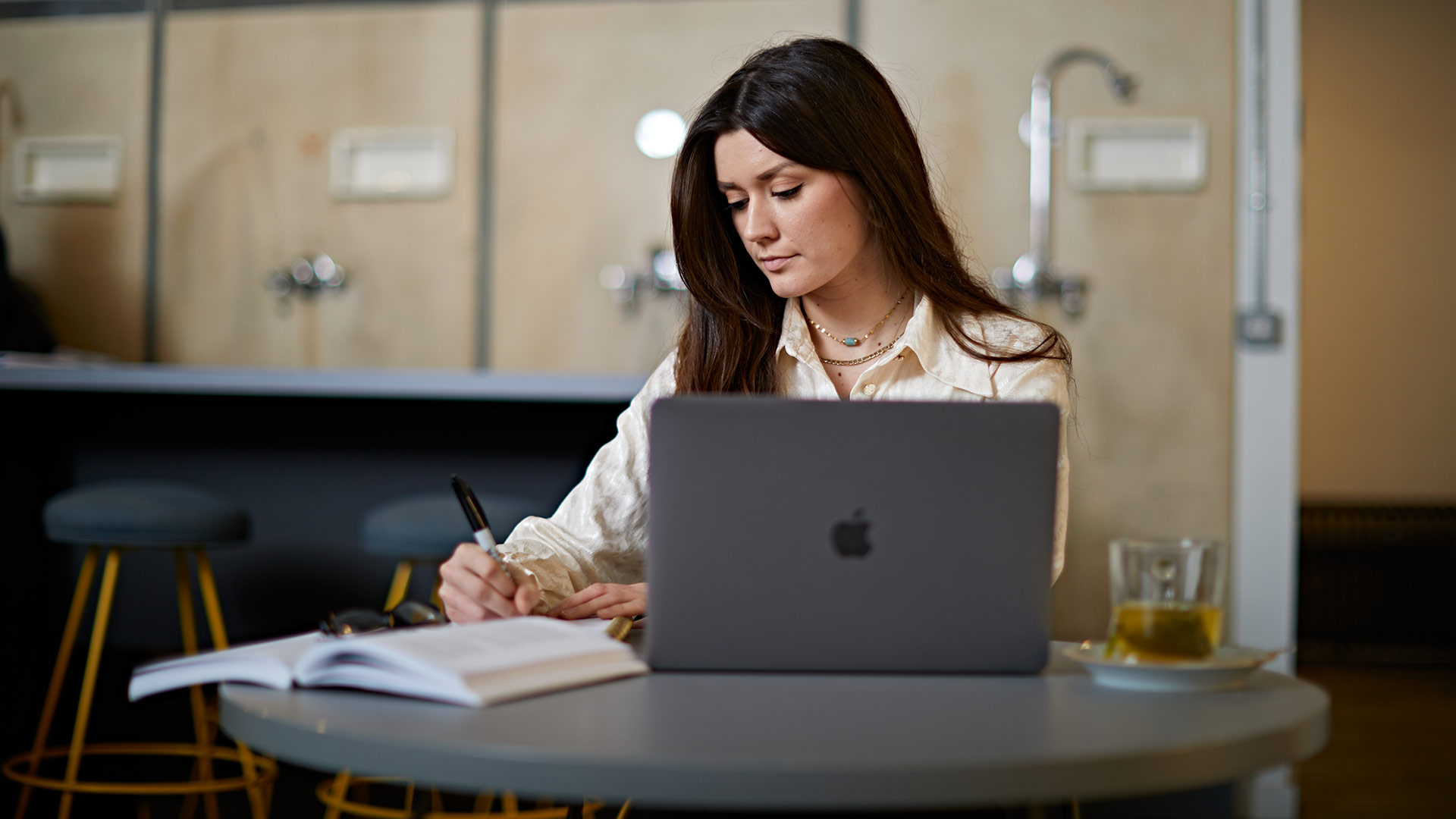 A student sat writing notes in a café, with their laptop in front of them on the table.