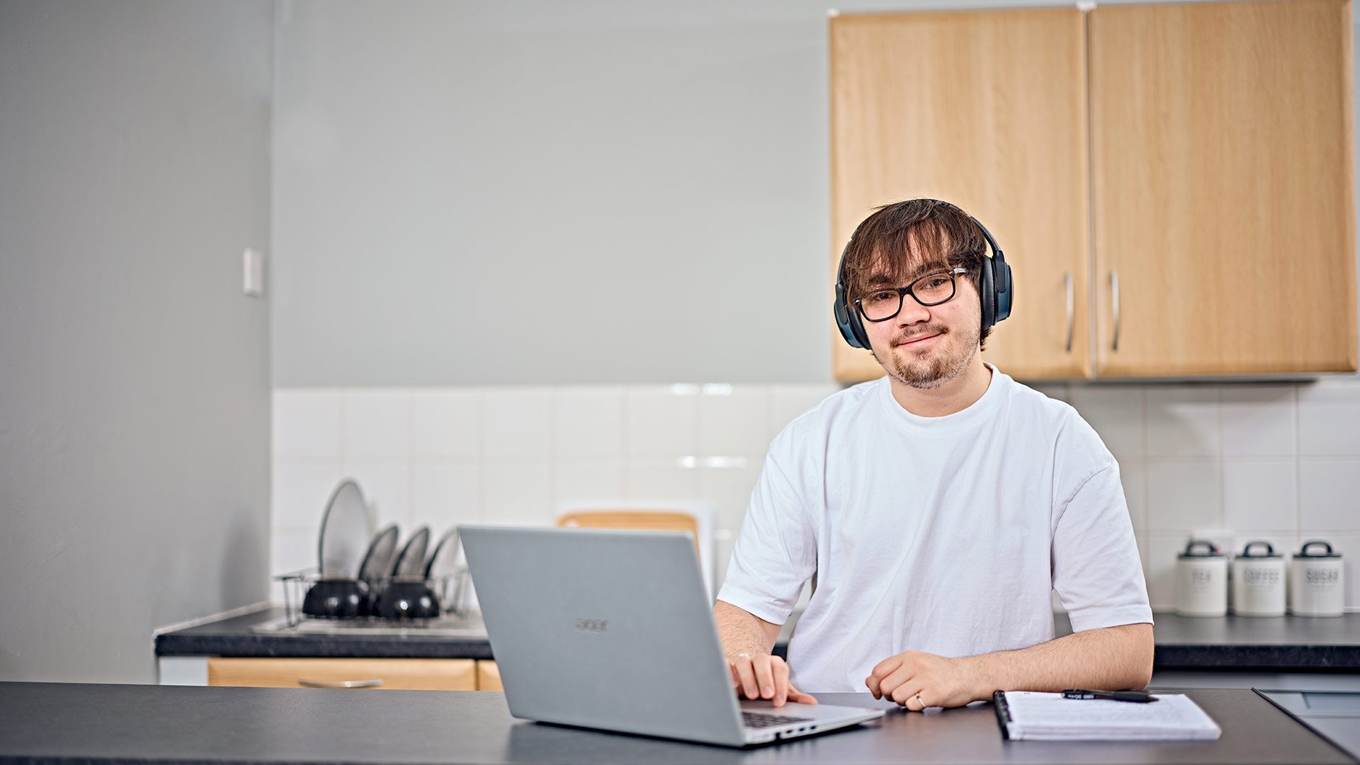 A student wearing a headset, working on a laptop in their kitchen.