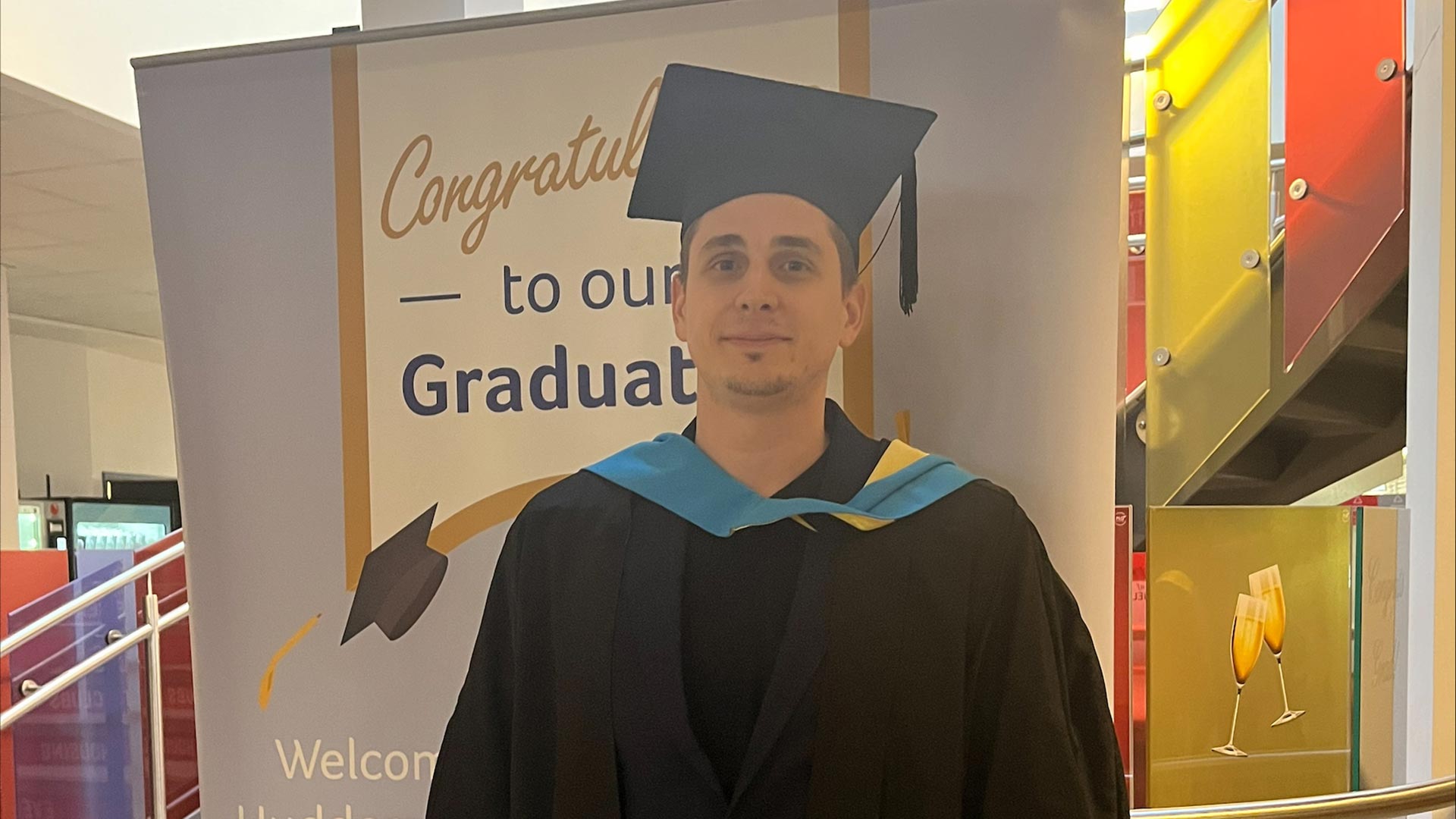 Stefano stood in front of a graduation pull up banner. He is wearing a graduation hat and gown.