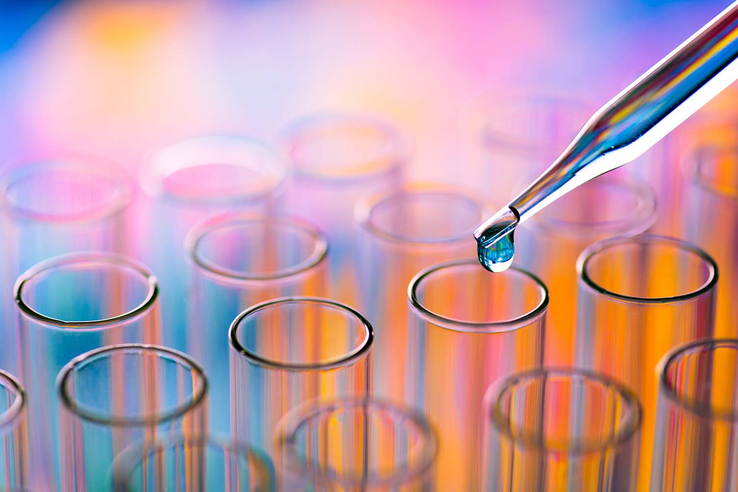 Close up photograph of a pipette dropping a clear liquid into a test tube, lit in orange and blue