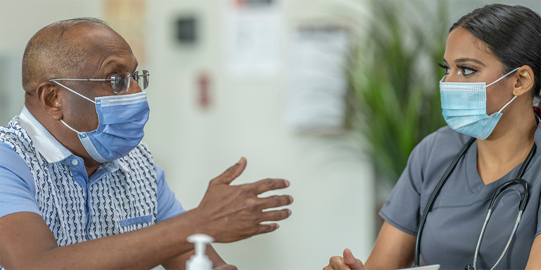 Close cropped image of a man wearing glasses talking to a young woman wearing a stethoscope. Both are wearing face masks.