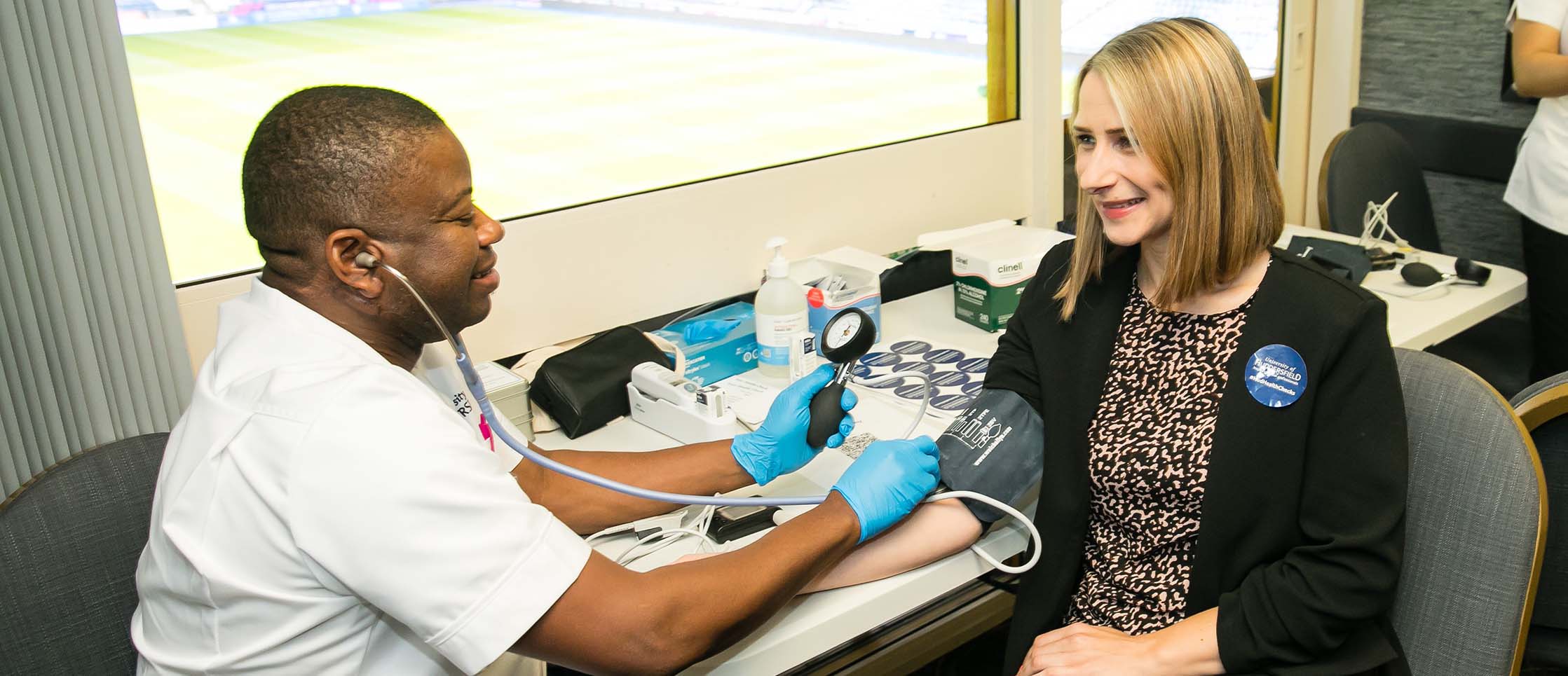 Two people are sitting opposite each other, the person on the left is taking the blood pressure of the person on the right. There is a large window next to them looking out onto a football pitch.