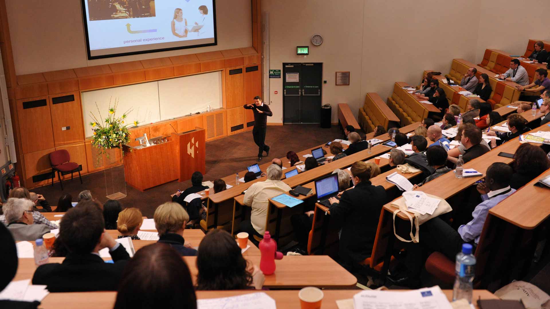 Many students receiving a lecture from a member of staff in a brown and cream lecture hall.