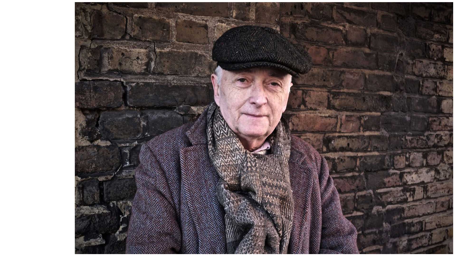 Christopher Reid the poet with a flat cap leaning against a brick wall