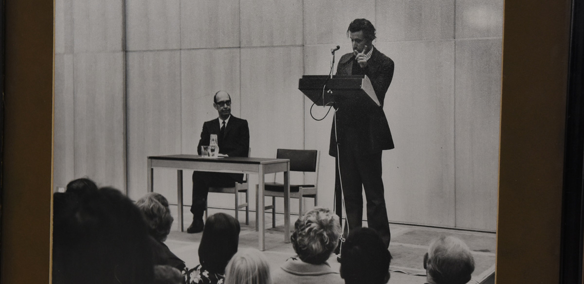 Ted Hughes speaking at the University of Hull with a disconsolate Philip Larkin sat at the desk.  As librarian at the University, it is likely that Larkin was asked to attend and host the event.
