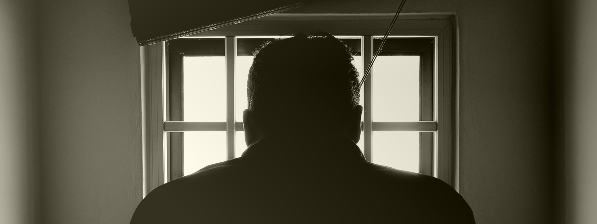 A sepia coloured photo of the back of a man looking outside of a window in prison