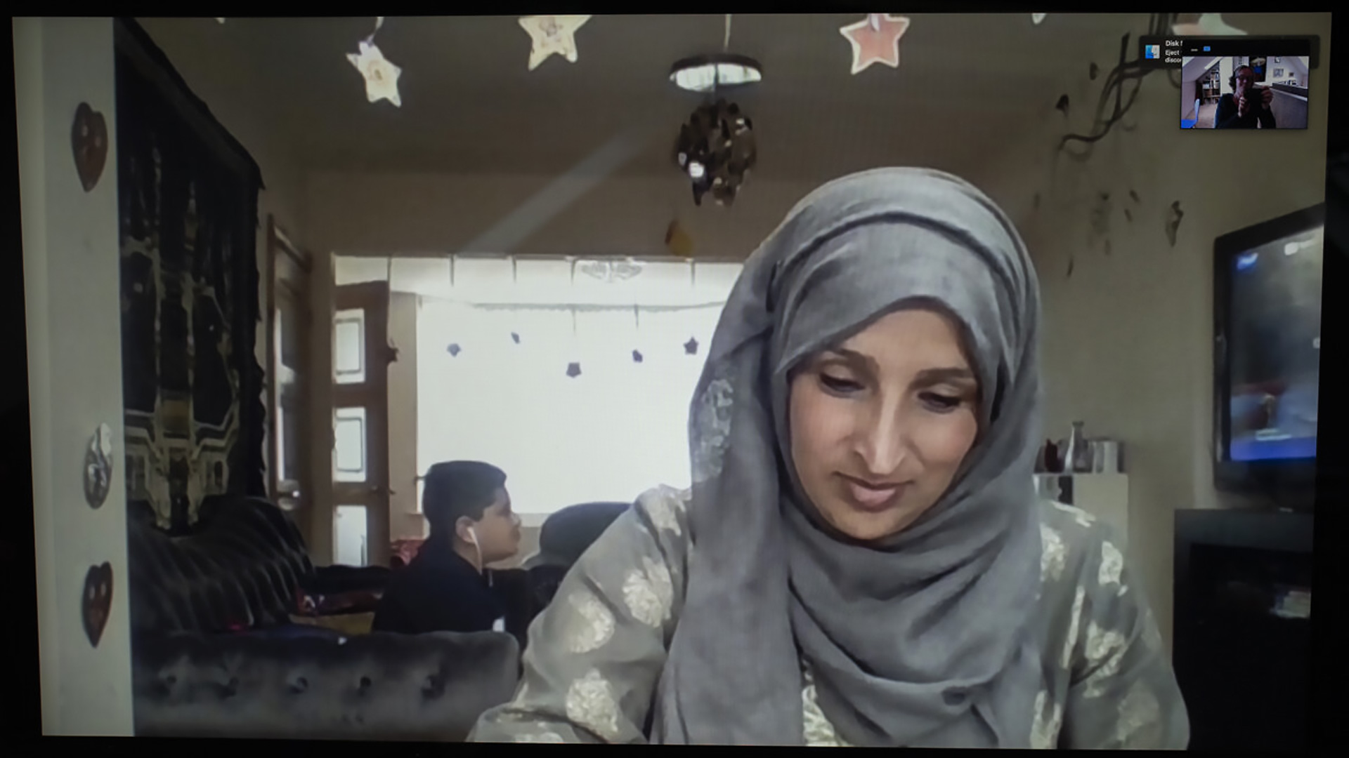 Shabnam sits in the foreground whilst her youngest child uses the games console behind her to communicate with friends whilst she works.