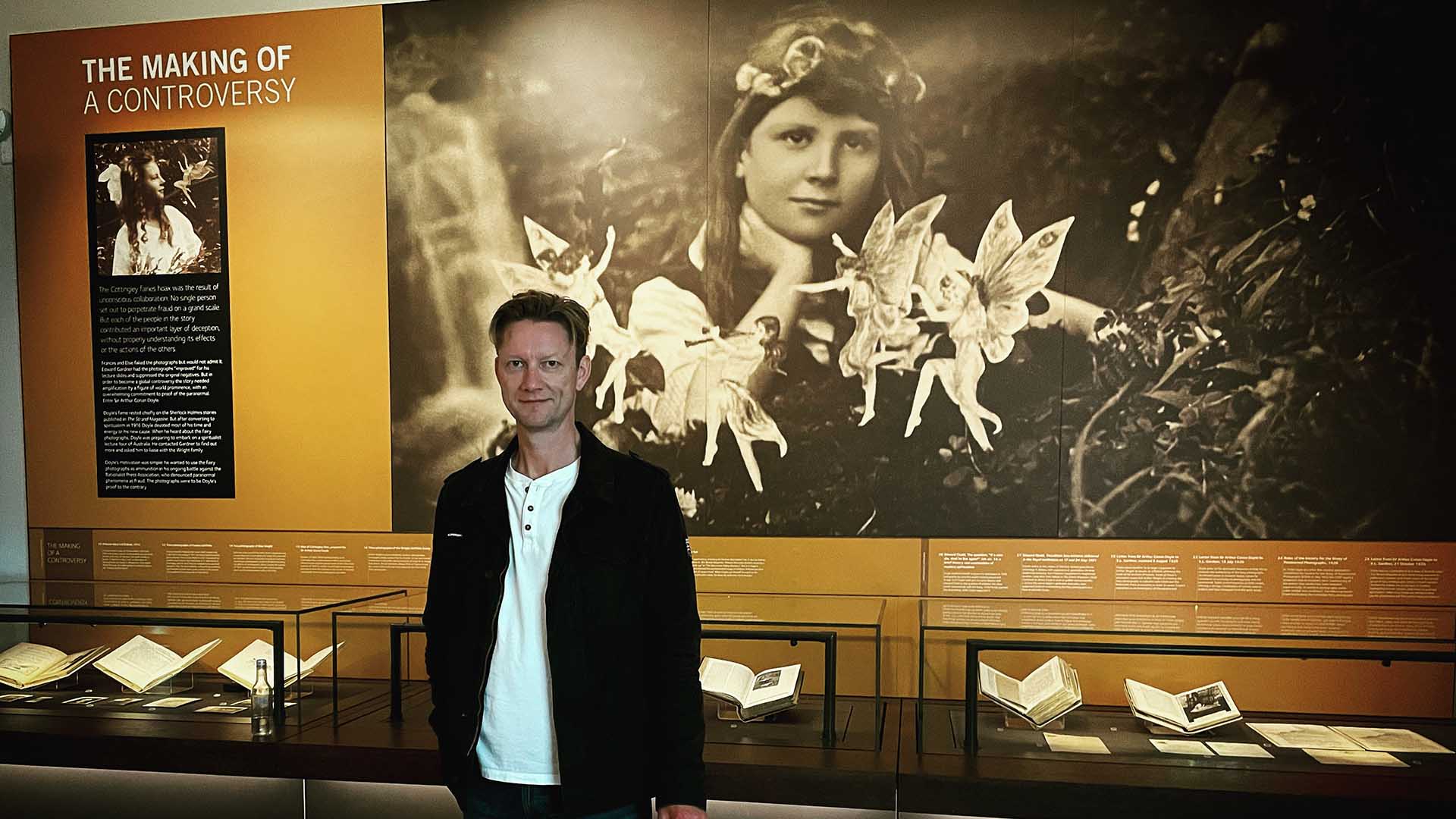Dr Merrick Burrow stands in front of a photo of the Cottingley fairies