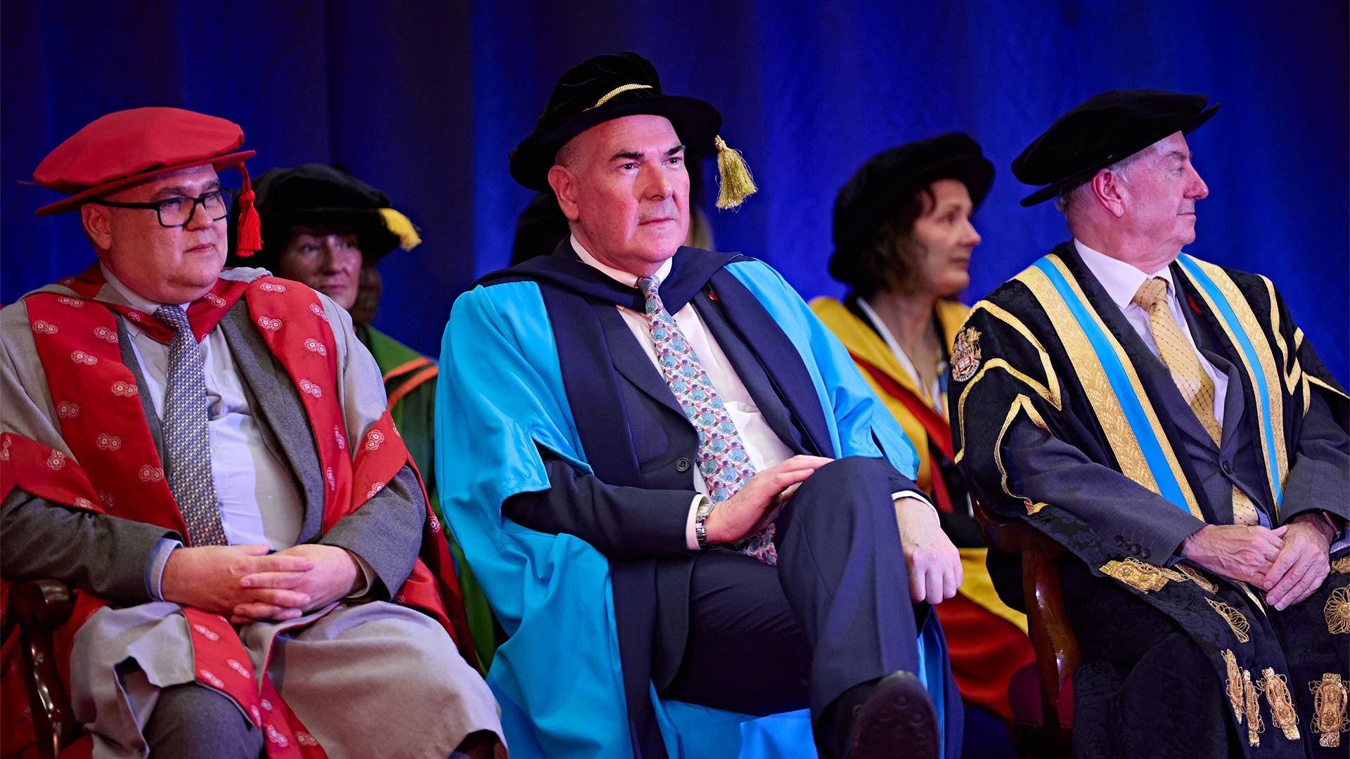 A man sat on stage wearing a graduation gown during a ceremony