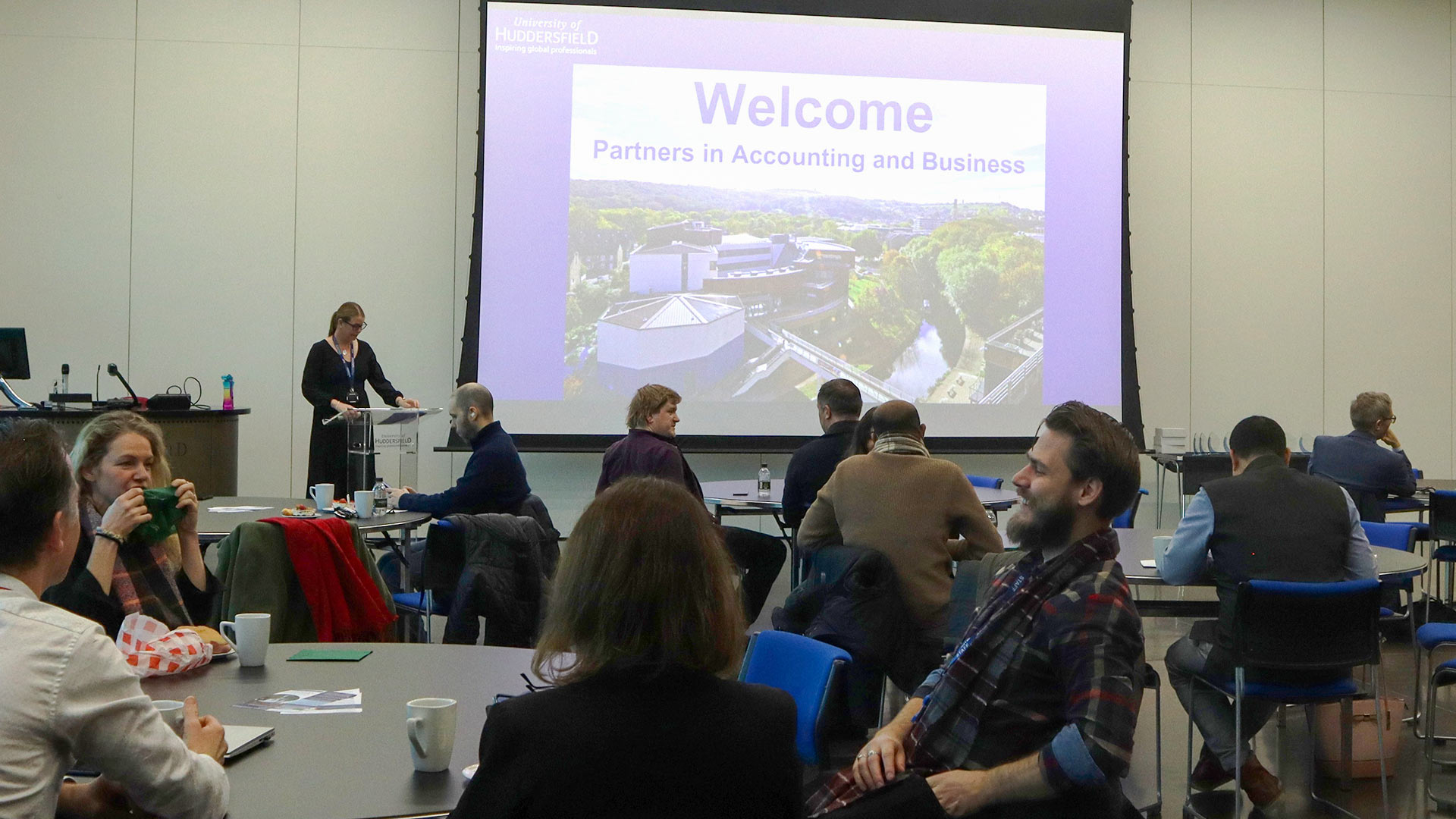 A group of people sat in a conference room facing a screen with one person presenting