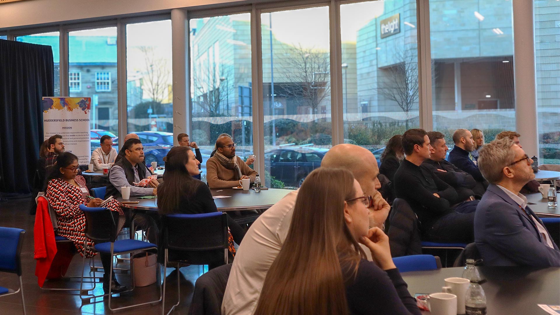 A group of people sat in a conference room facing a screen with one person presenting