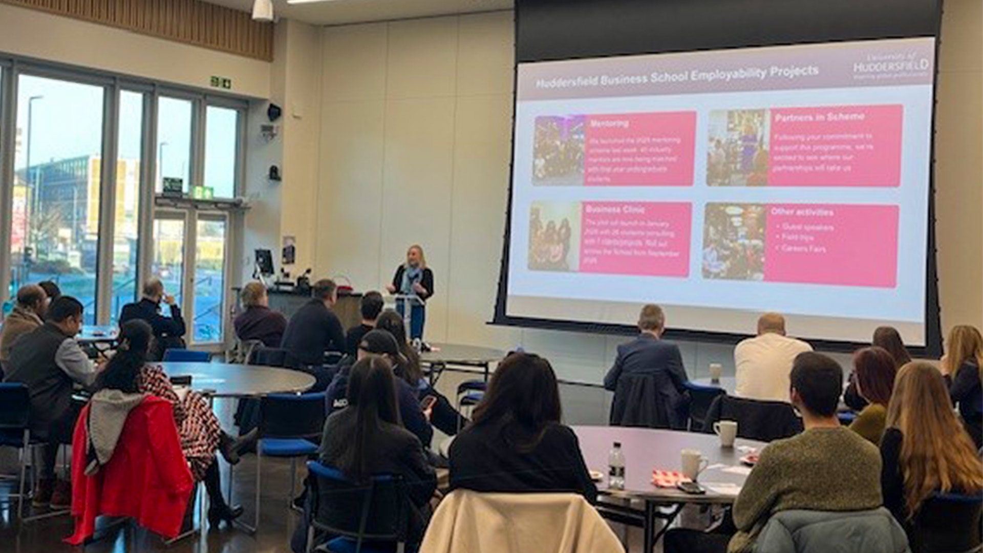 A group of people sat in a conference room facing a screen with one person presenting