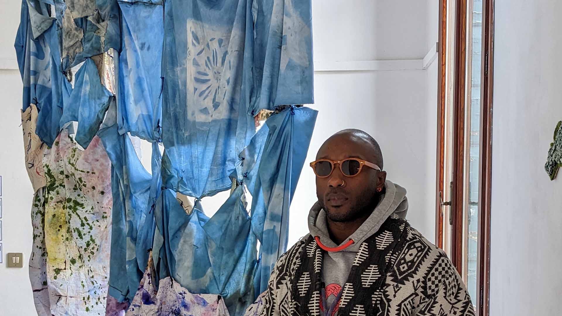 Benaiah Matheson sits in a gallery with his art work wearing sunglasses