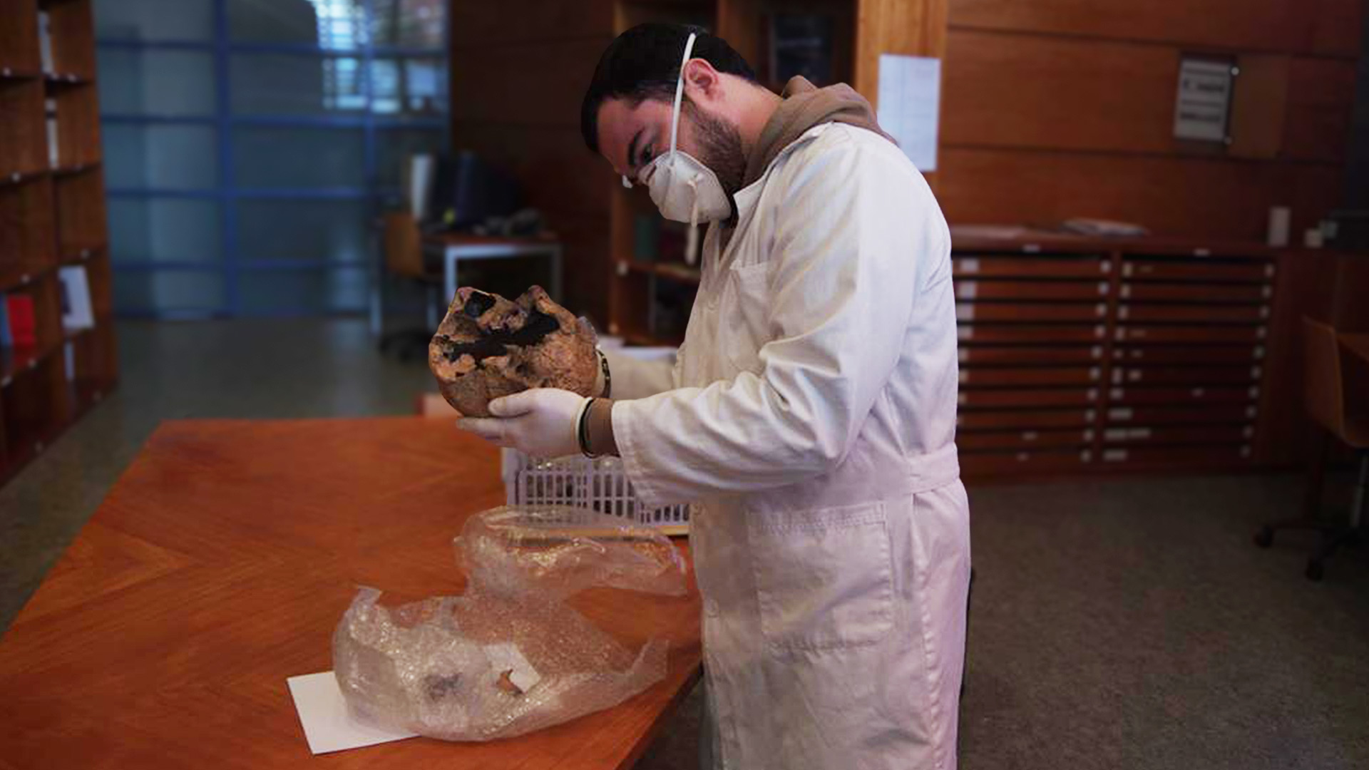 A man in a protective suit and mask studying a skull in a lab