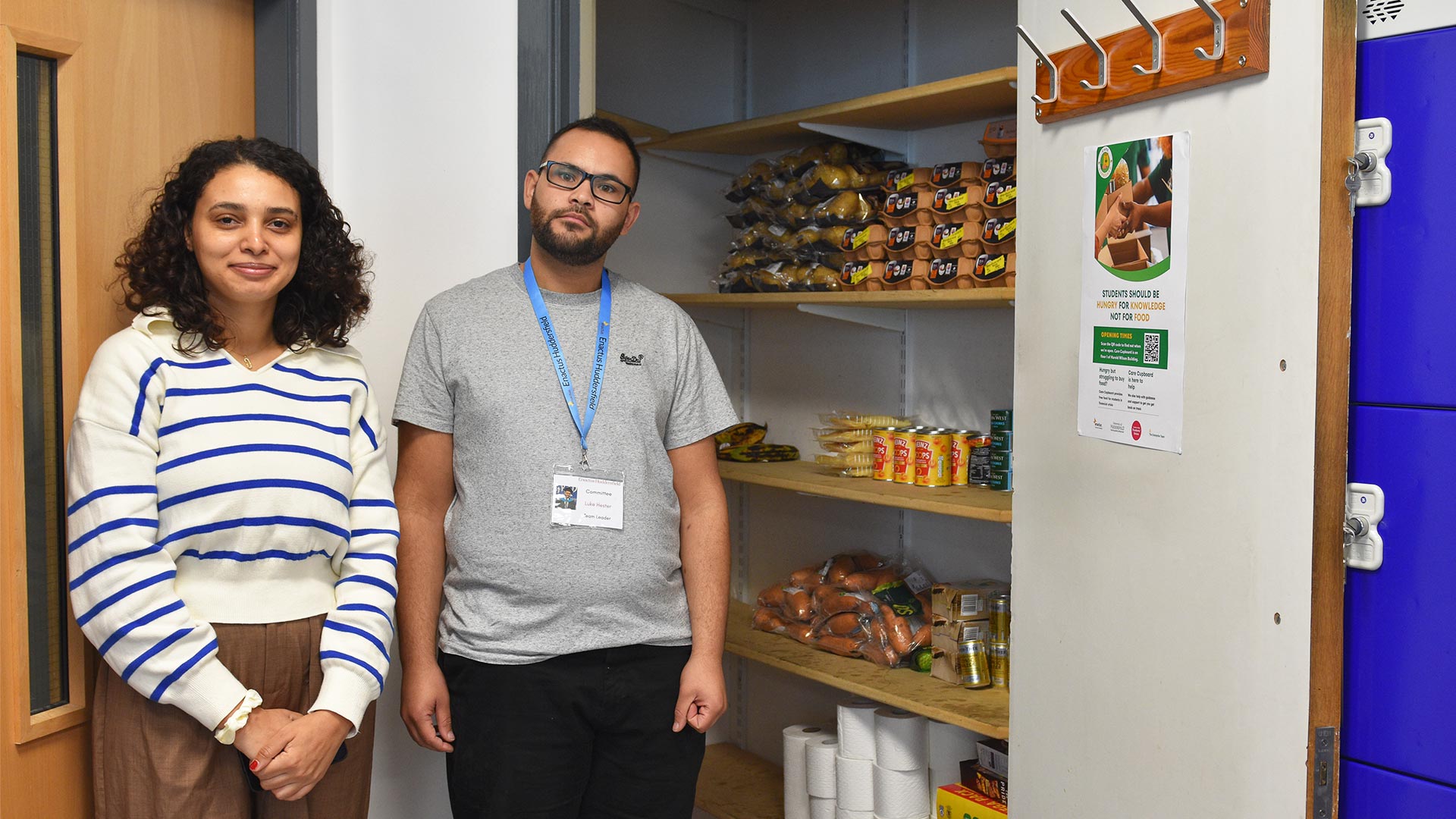 Two students stood next to the open door of a cupboard with shelves stocked with food items