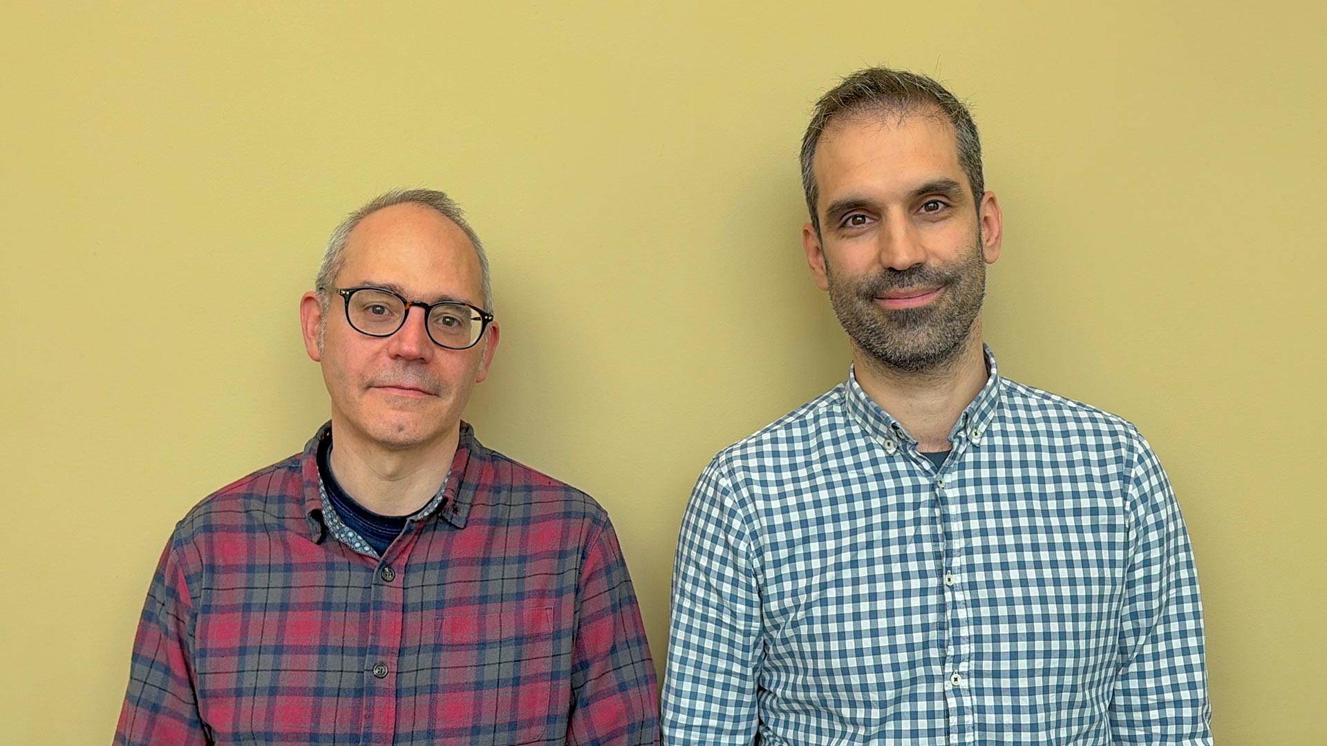 Two people standing side by side against a plain yellow wall, wearing casual long-sleeved shirts, facing the camera.