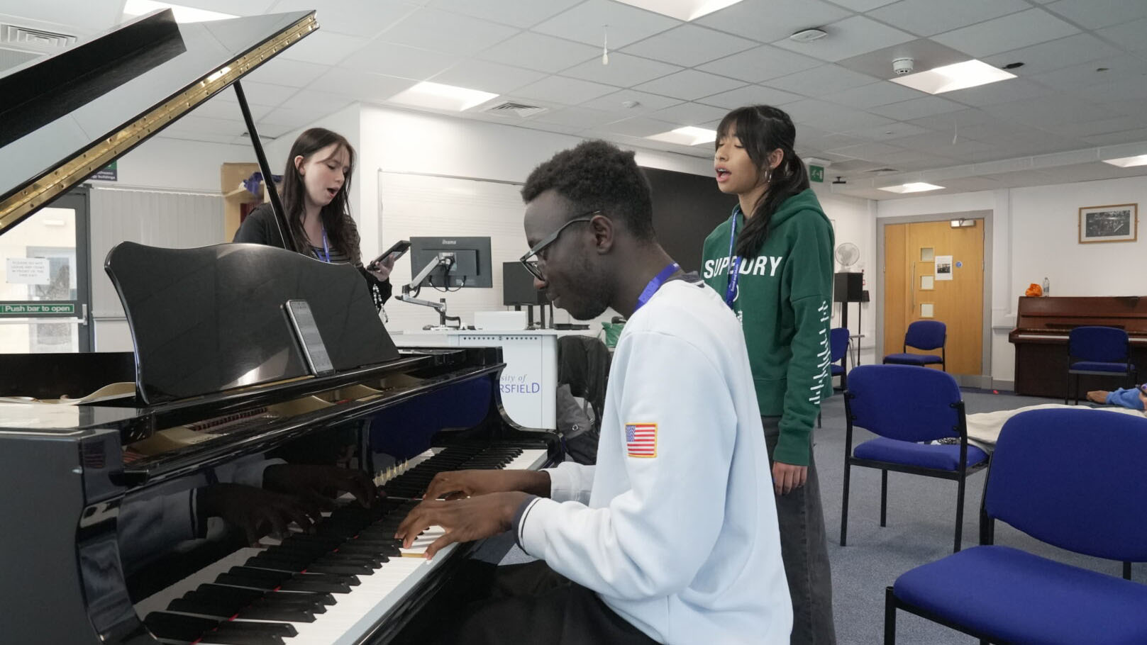 A young man plays a piano while two young women sing near him in a room at the University of Huddersfield