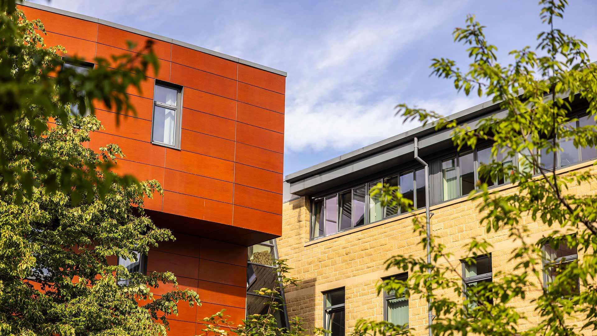 University campus buildings framed by trees, featuring orange cladding and stone façades, viewed against a blue sky with light cloud cover.