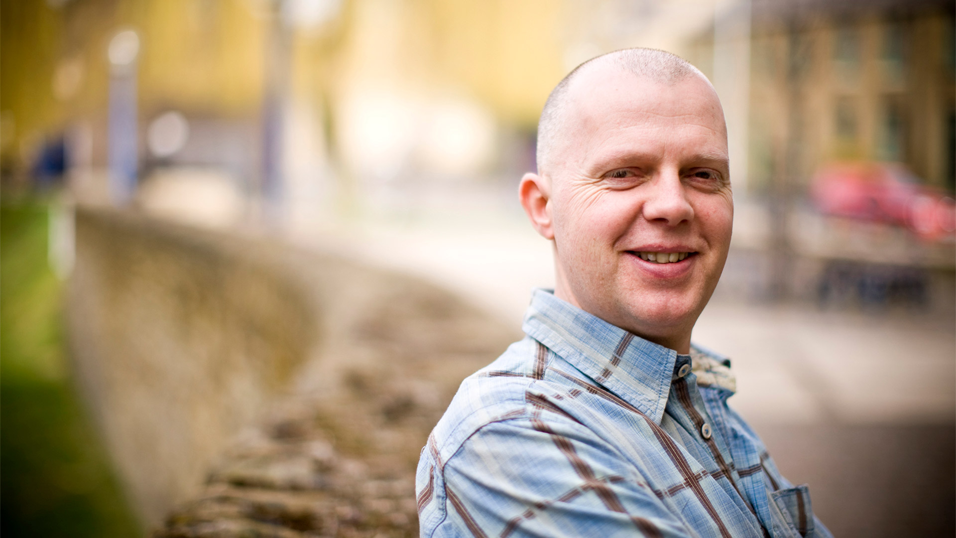 Outdoor portrait of an individual in a checked shirt standing beside a stone wall, with a softly blurred streetscape and buildings visible in the background.