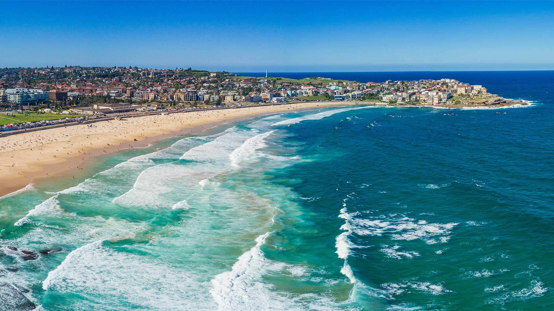 Aerial view of Bondi Beach coastline, showing sandy beach, turquoise ocean waves, surrounding residential areas and headland extending into the Pacific Ocean.