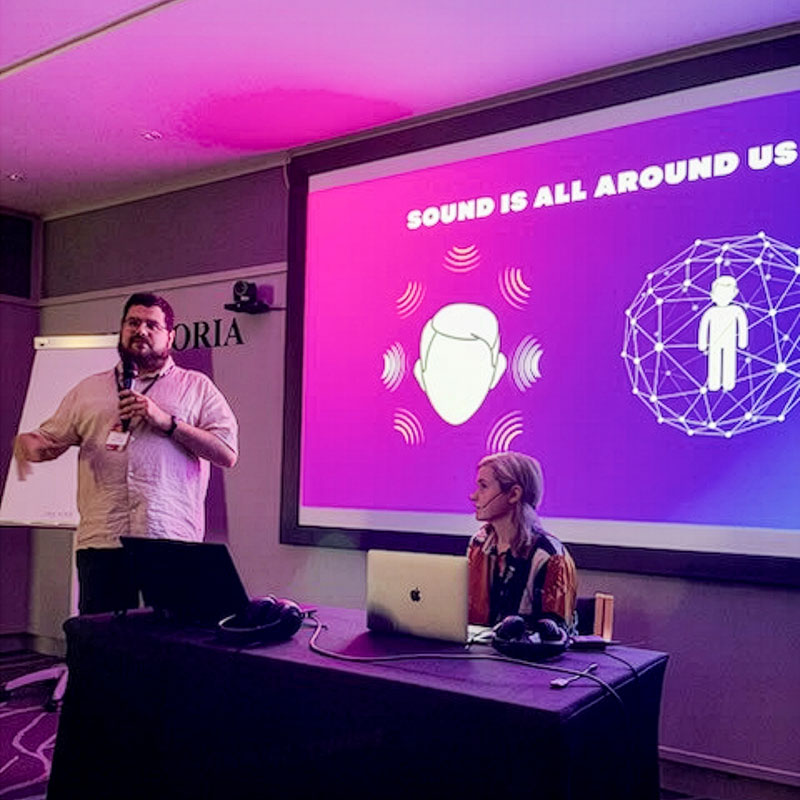 A bearded man in a light pink short sleeved shirt stands on the left of a conference room speaking into a microphone, while a woman with light hair, wearing a patterned top, sits at a table on the right with an open laptop and headphones. Behind them, a large presentation screen displays the text “Sound is all around us” on a purple background with graphics showing sound waves