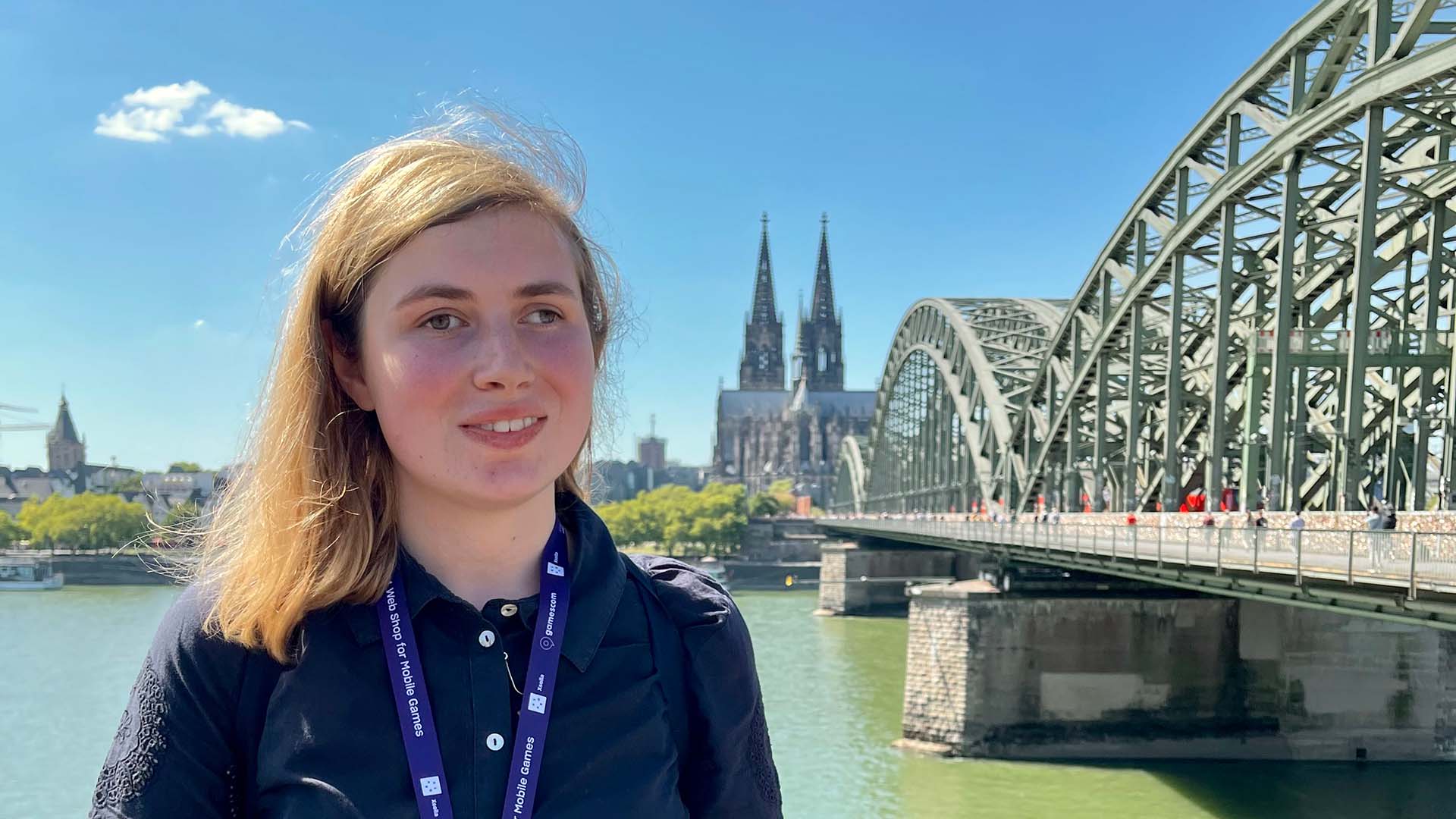 A young woman with light, shoulder length hair stands by a river on a sunny day. She is wearing a dark, short sleeved shirt and a DevCom lanyard with a badge, holding a folded cane in front of her. Behind her, a large green steel arch bridge crosses the river, with a city skyline and twin cathedral spires visible in the distance beneath a clear blue sky.