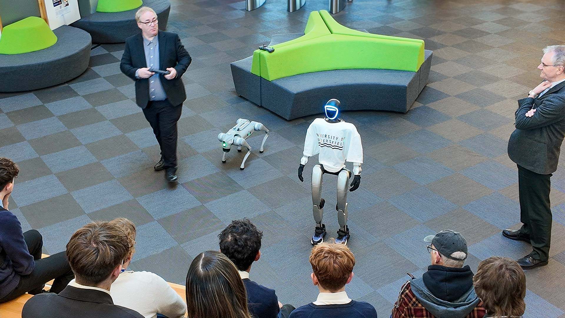 A man demonstrating humanoid and dog robots in front of a group of students