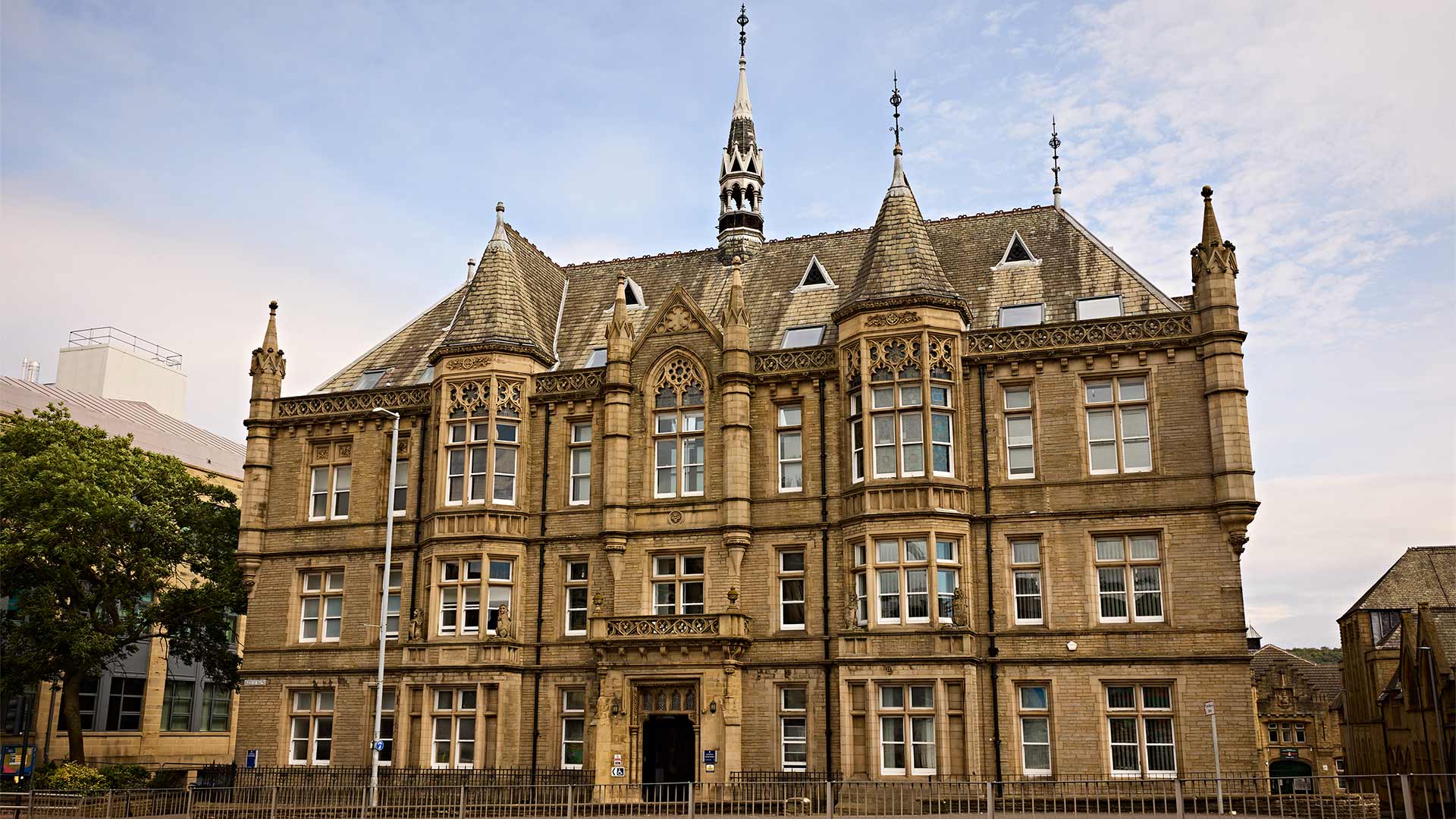 Historic Gothic-style university building with ornate stone façade, towers and spire, viewed from across the road under a partly cloudy sky.