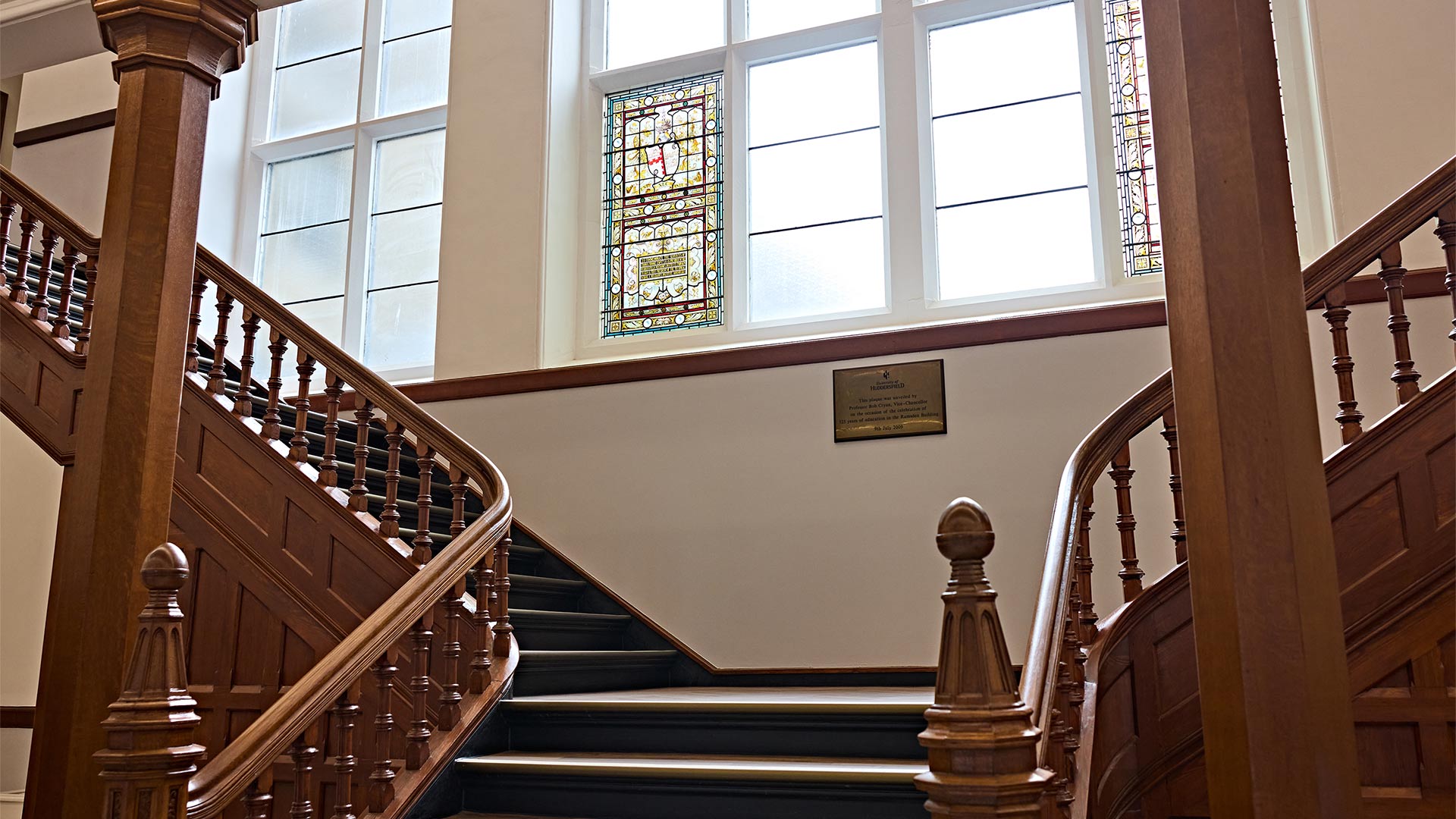 Grand wooden staircase inside a restored historic building, featuring stained-glass windows, decorative balustrades and original architectural details.