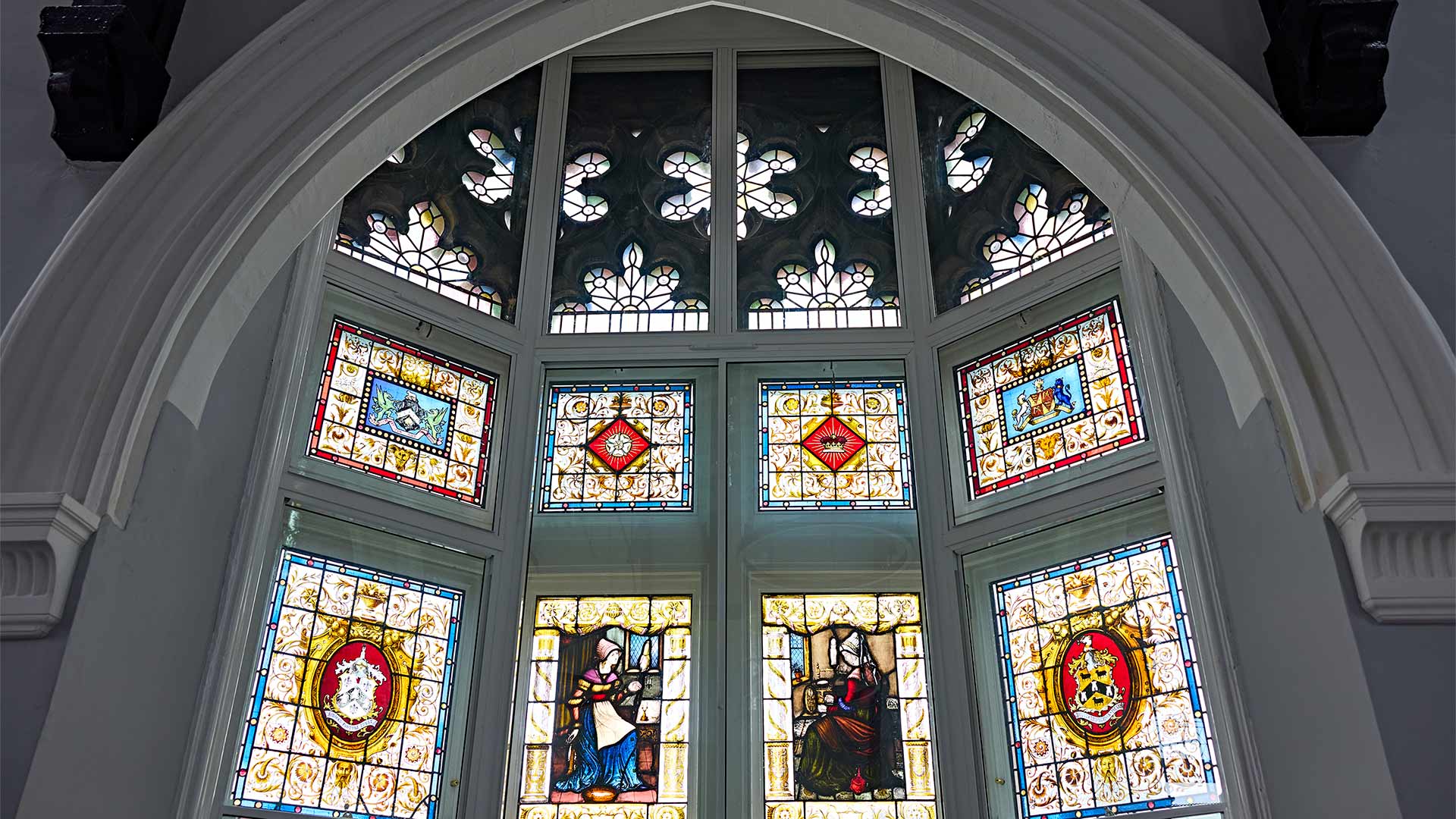Ornate stained-glass window beneath Gothic archway, featuring decorative patterns, heraldic emblems and figurative panels preserved within a restored historic building.