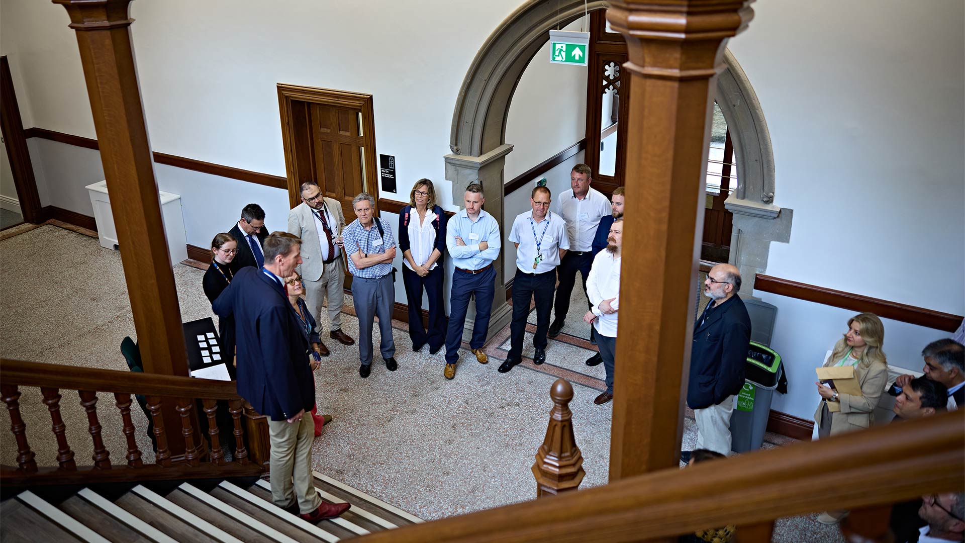 Visitors stand in an entrance hall with stone archways, wooden staircases and original architectural features during a guided building tour.