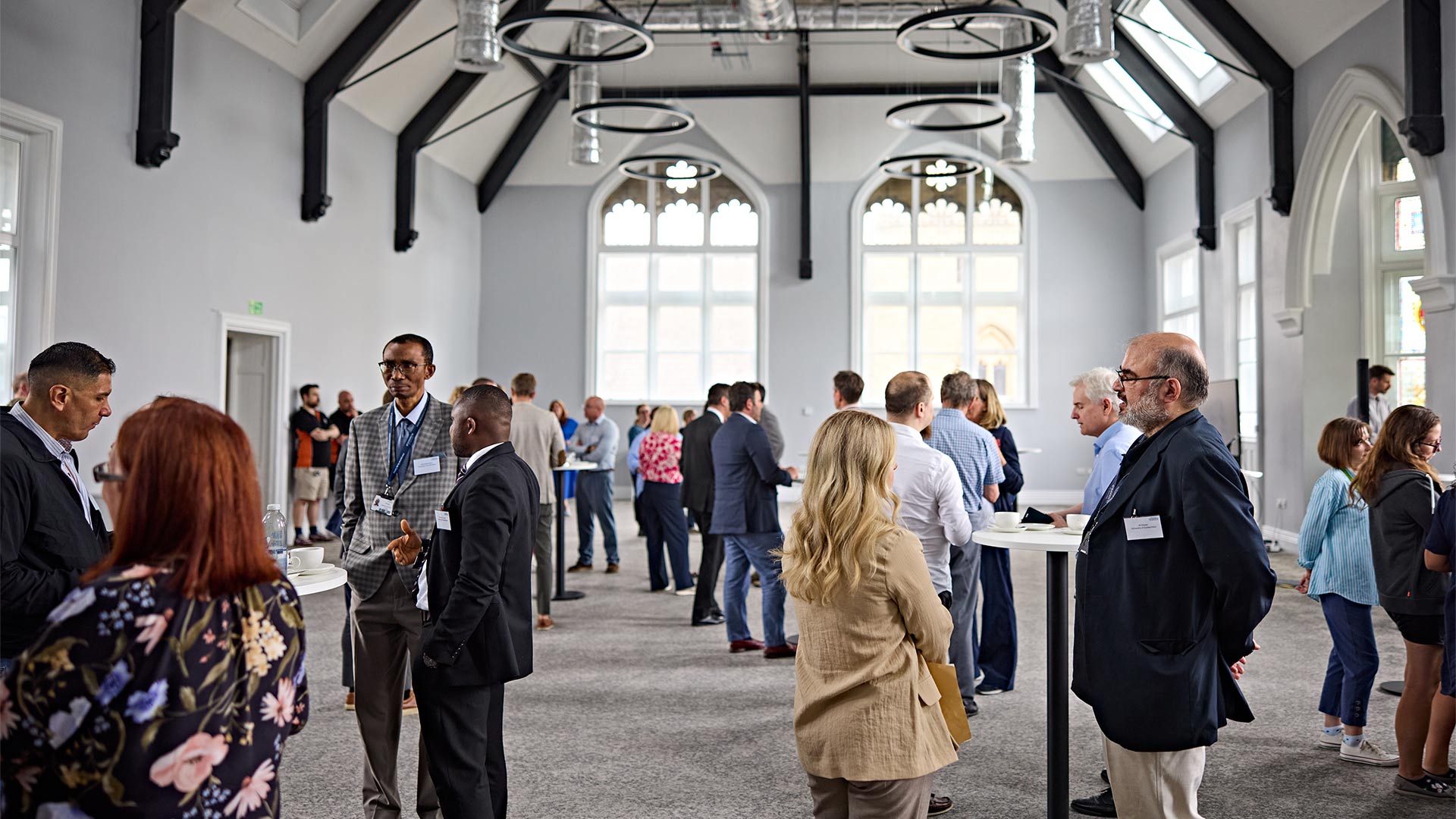 Guests network in a large restored hall with high ceilings, arched windows and exposed beams during an event inside a historic building.