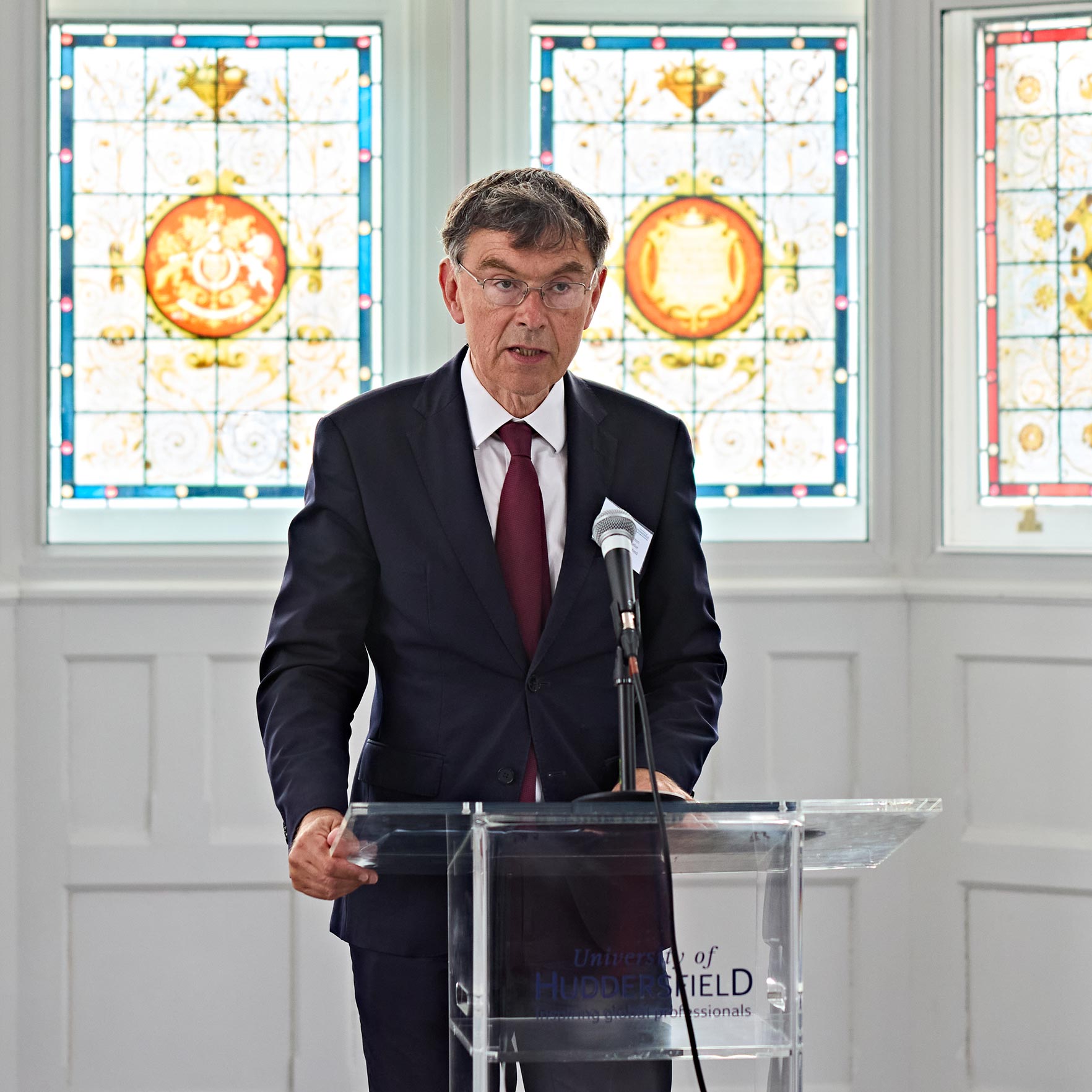 Speaker stands at a lectern in front of stained-glass windows, delivering remarks during an event in a restored historic building.