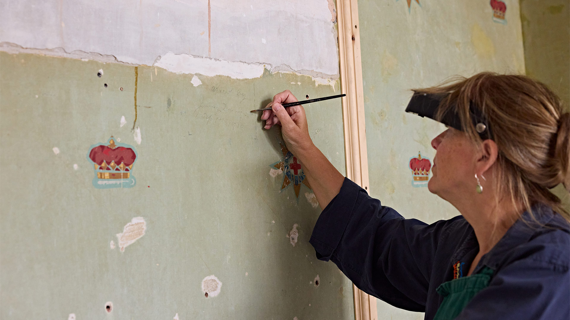 Conservation specialist examines historic wallpaper on a wall, revealing preserved heritage features during restoration work inside a historic building.