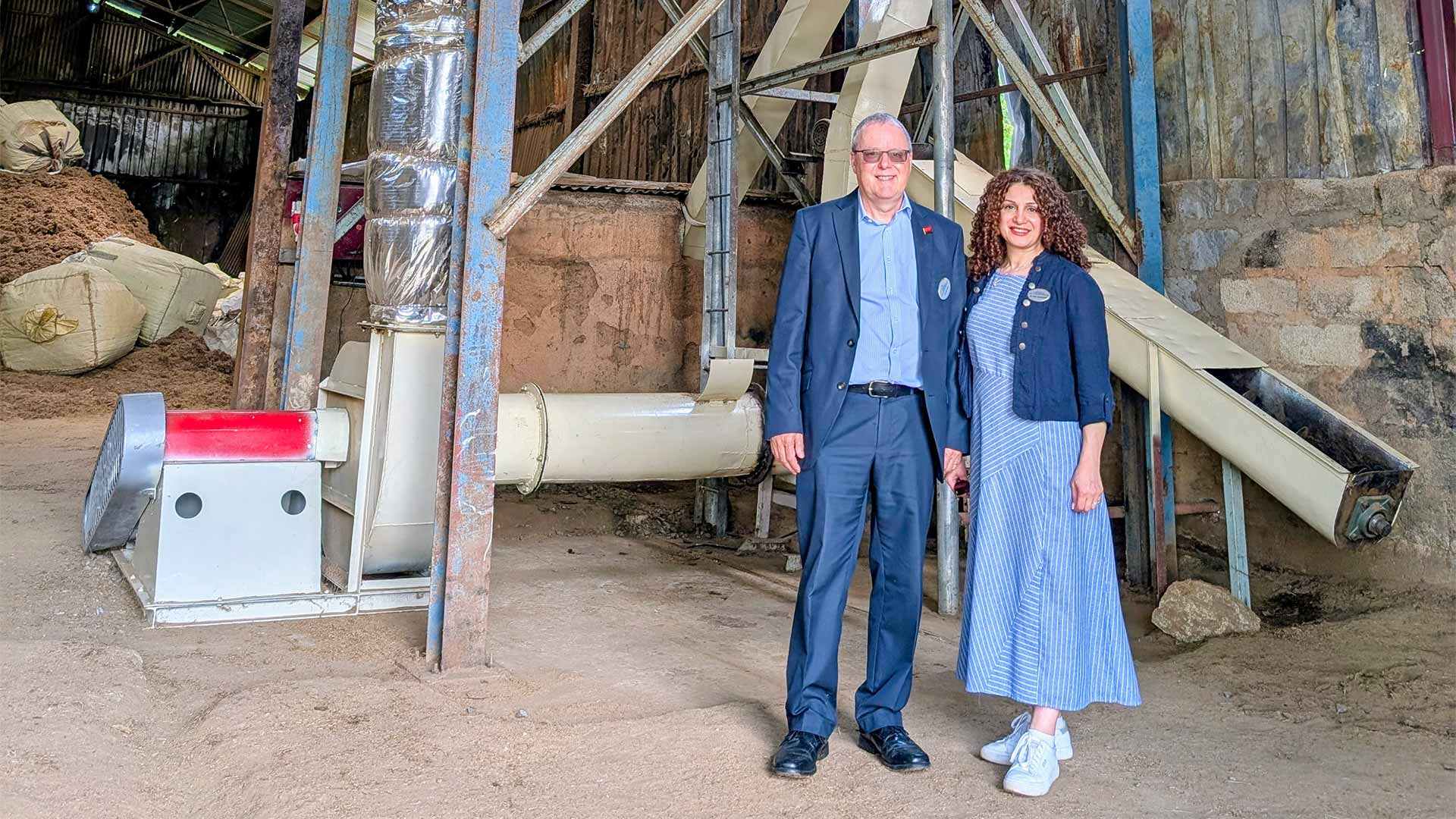 Two individuals stand inside industrial processing facility beside large machinery and ducting, with piles of raw material and metal framework visible in a warehouse setting.