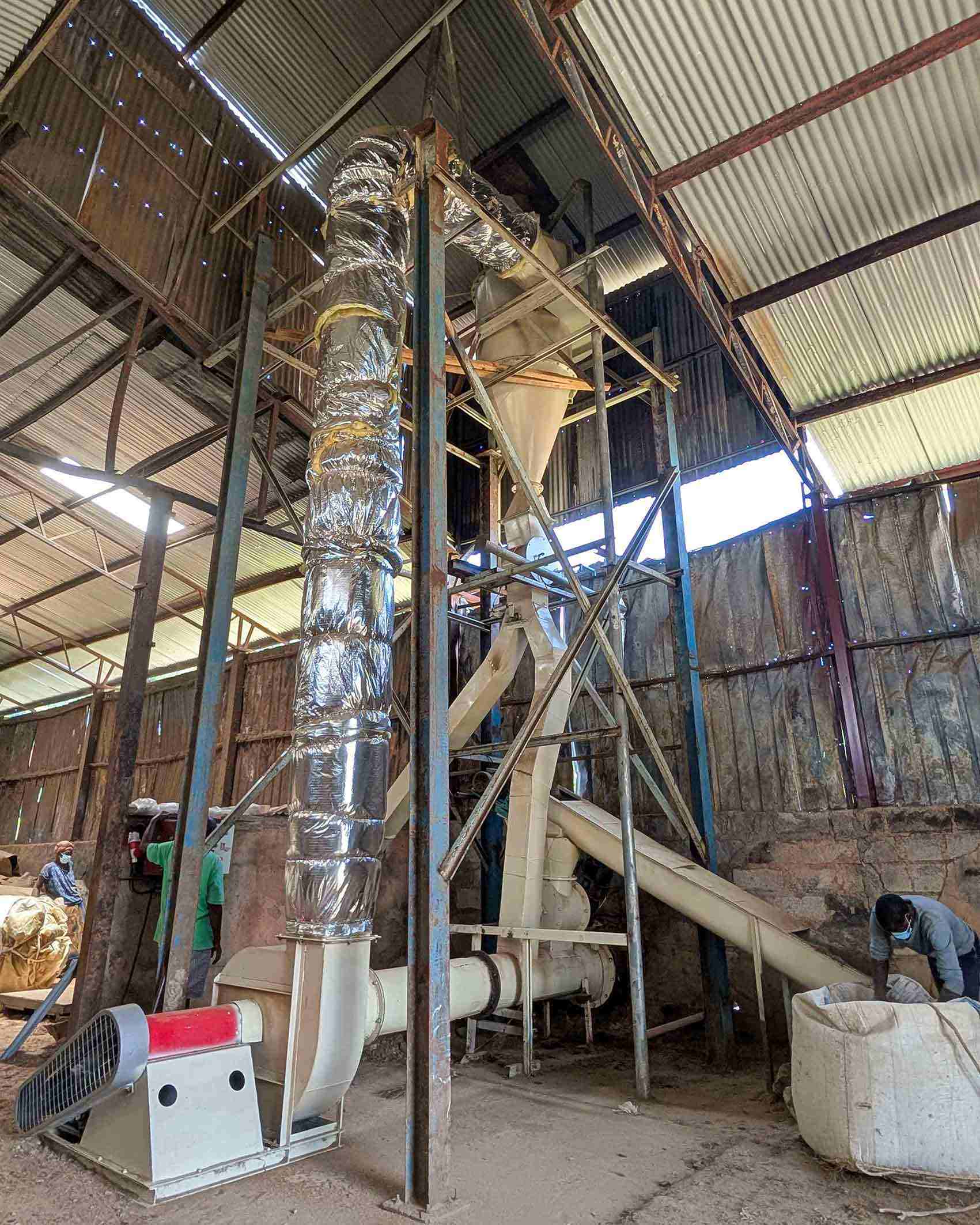 Interior of industrial building showing tall processing equipment, enclosed piping and metal framework, with large sacks and workers operating within the space.