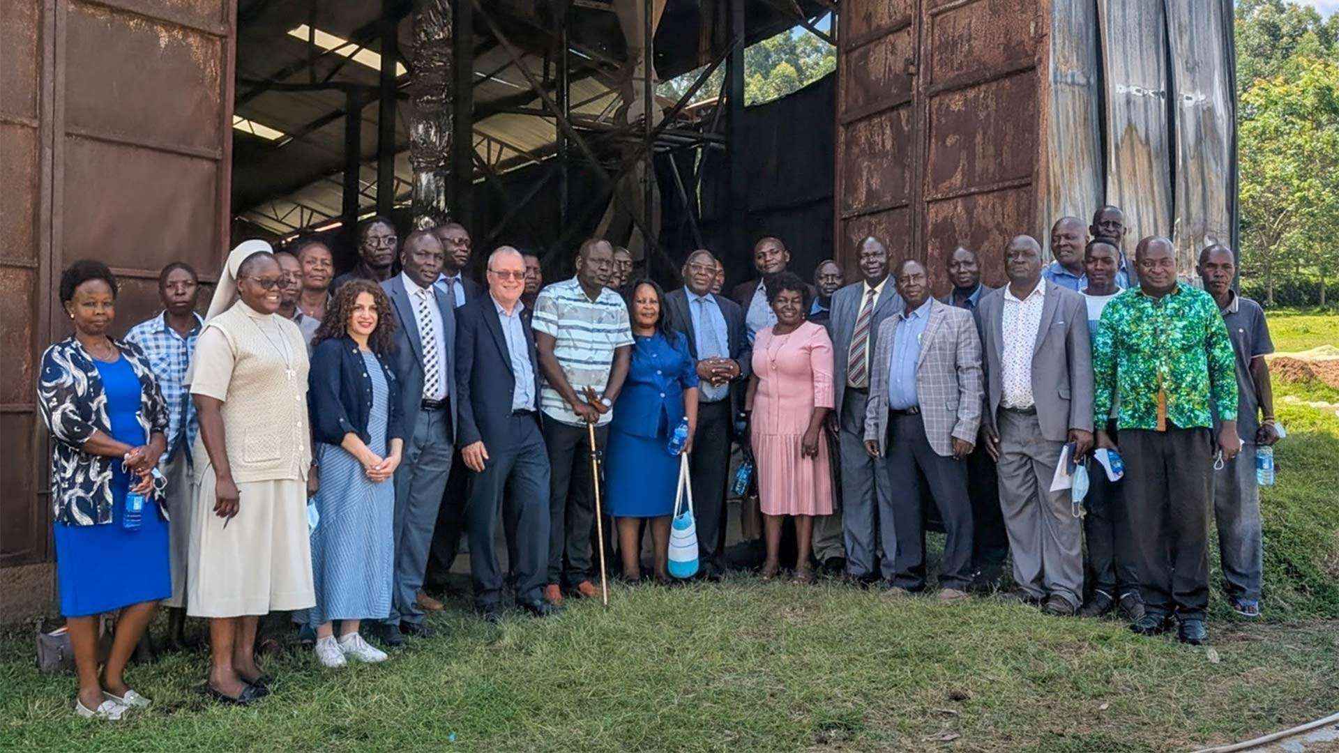 Large group stands outside open industrial building entrance, gathered for group photo, with machinery visible inside and grassy area in the foreground.