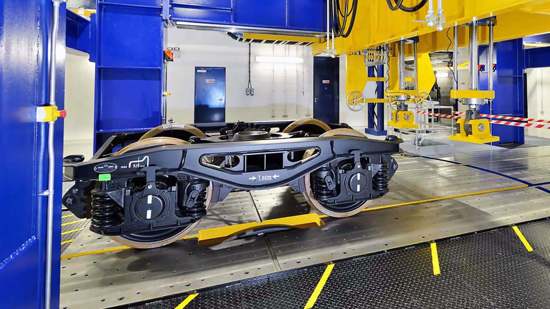 Railway bogie mounted on testing rig inside engineering laboratory, with blue and yellow machinery and industrial equipment visible.