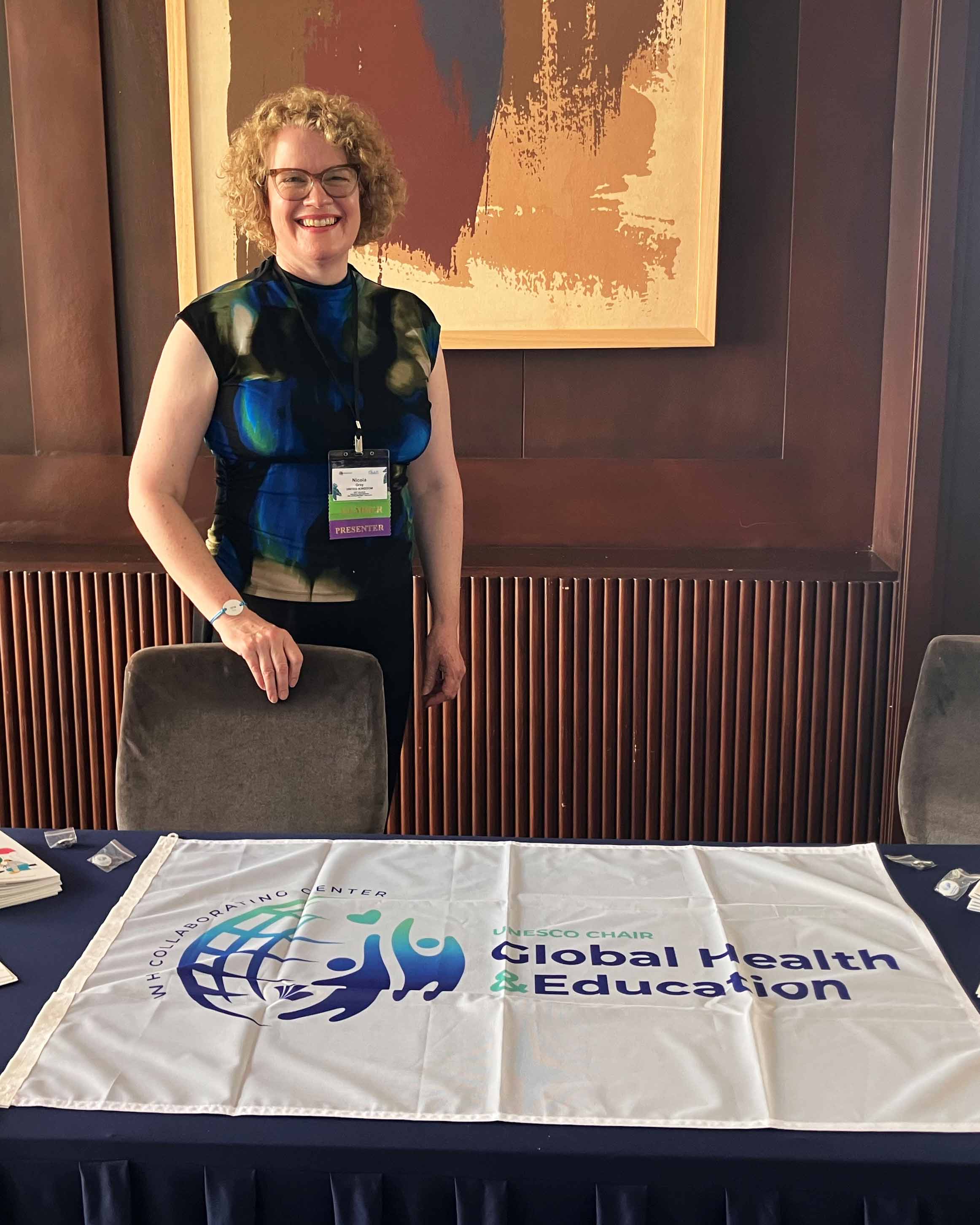 Individual wearing conference badge stands behind table displaying UNESCO Chair Global Health and Education banner, with printed materials and abstract artwork on wall behind.